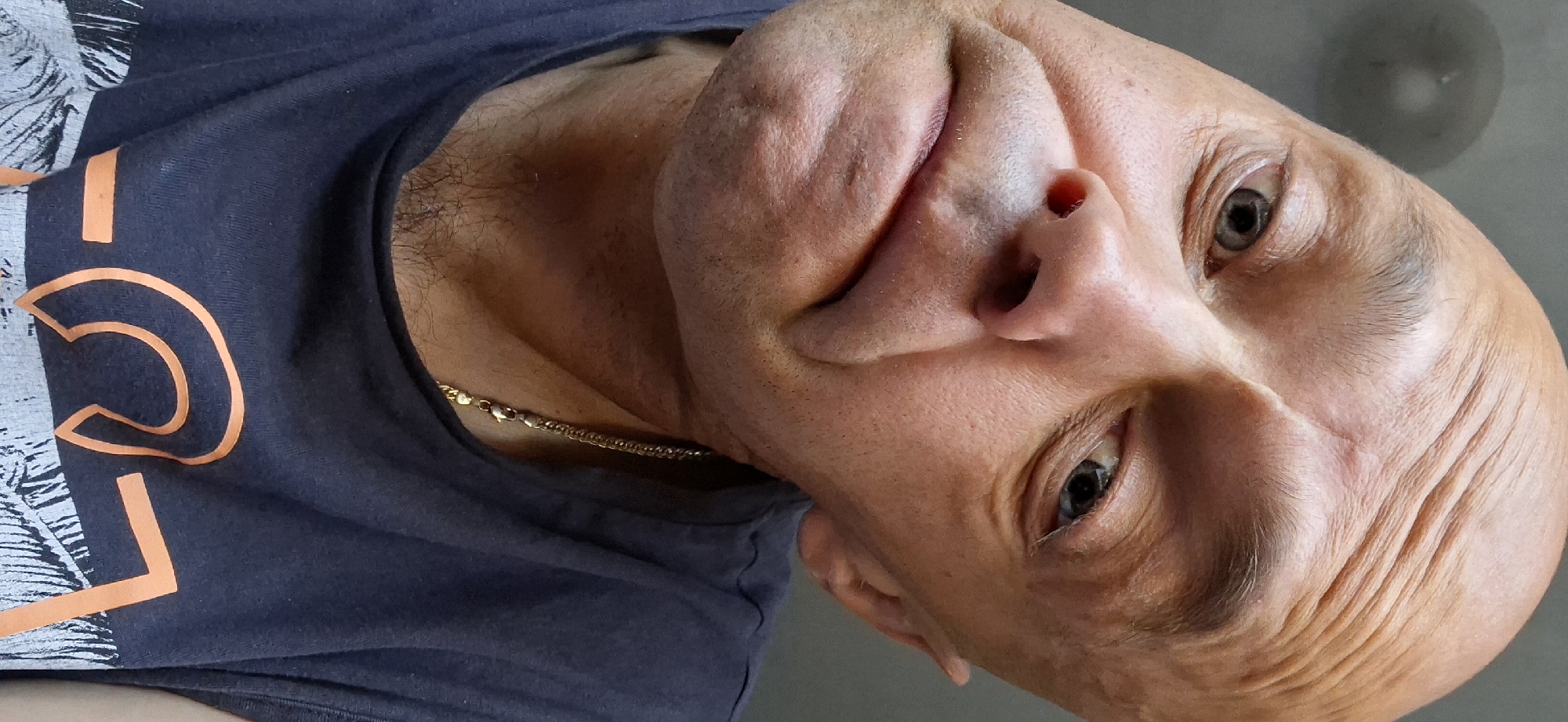 The image is a close-up, low-angle selfie of a bald man with light skin and visible wrinkles. He is wearing a dark blue t-shirt with an orange graphic design and a gold chain necklace. The man's head is tilted, and he has a slightly distorted facial expression with his mouth turned downwards. His eyes are open, and one is looking towards the camera. The background is a plain, neutral gray, and there is a circular, blurry object in the upper left corner, possibly a light fixture. The scene appears to be indoors. There is no indication of the specific location within the city of Kamianets-Podilskyi, Ukraine.