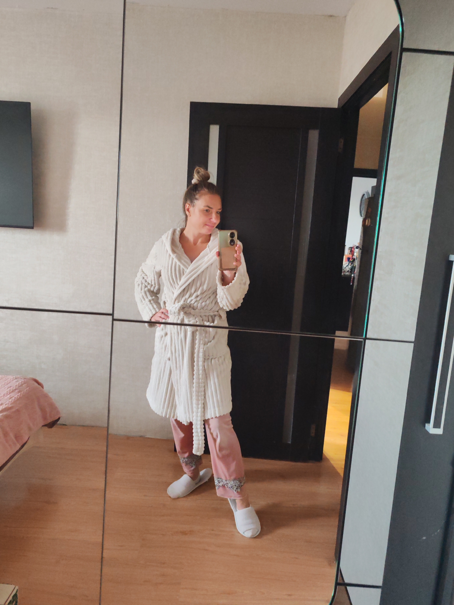 A woman captures a mirror selfie indoors, likely within a bedroom or dressing area in Petropavlovsk-Kamchatsky, Russia. She stands casually, holding a light-colored smartphone in her right hand, while her left hand rests on her hip. Her hair is pulled up in a relaxed messy bun, and she sports a soft, pleasant smile.
She is dressed in comfortable loungewear, consisting of a cream-colored, textured bathrobe with a hood and a tie belt, worn over pink pajama bottoms featuring decorative gray cuffs near the ankles. White slippers complete her cozy attire.
The setting is a modern room characterized by light-colored walls and light wooden flooring. A large mirror, segmented by dark lines, serves as the reflective surface for the selfie, suggesting it's part of a wardrobe or closet. Reflected in the mirror, on the left, is a portion of a bed covered with a pink quilted blanket, and above it, a dark flat-screen television mounted on the wall. To the center-right, a dark wooden door frame with a slightly ajar door reveals a brightly lit area beyond. Further to the right, a dark gray or black closet panel with a minimalist white vertical handle is visible in the reflection. The overall scene conveys a relaxed, domestic atmosphere. No visible text is present in the image.