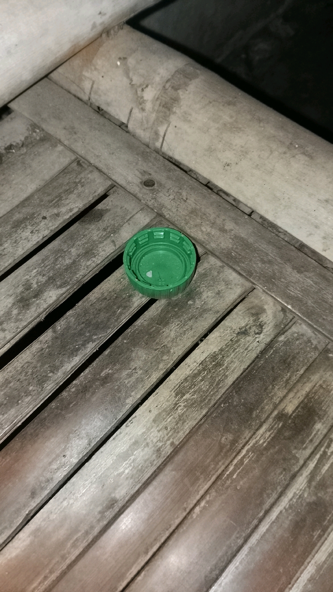 A green plastic bottle cap is resting on a weathered wooden surface, possibly a table or bench. The wood is laid out in parallel slats, showing signs of age and wear. The bottle cap is open and empty, with a small white speck visible inside. The background is dark and out of focus, suggesting it is an outdoor setting at night or in low light conditions. The overall scene is simple and focuses on the single object on the textured surface.