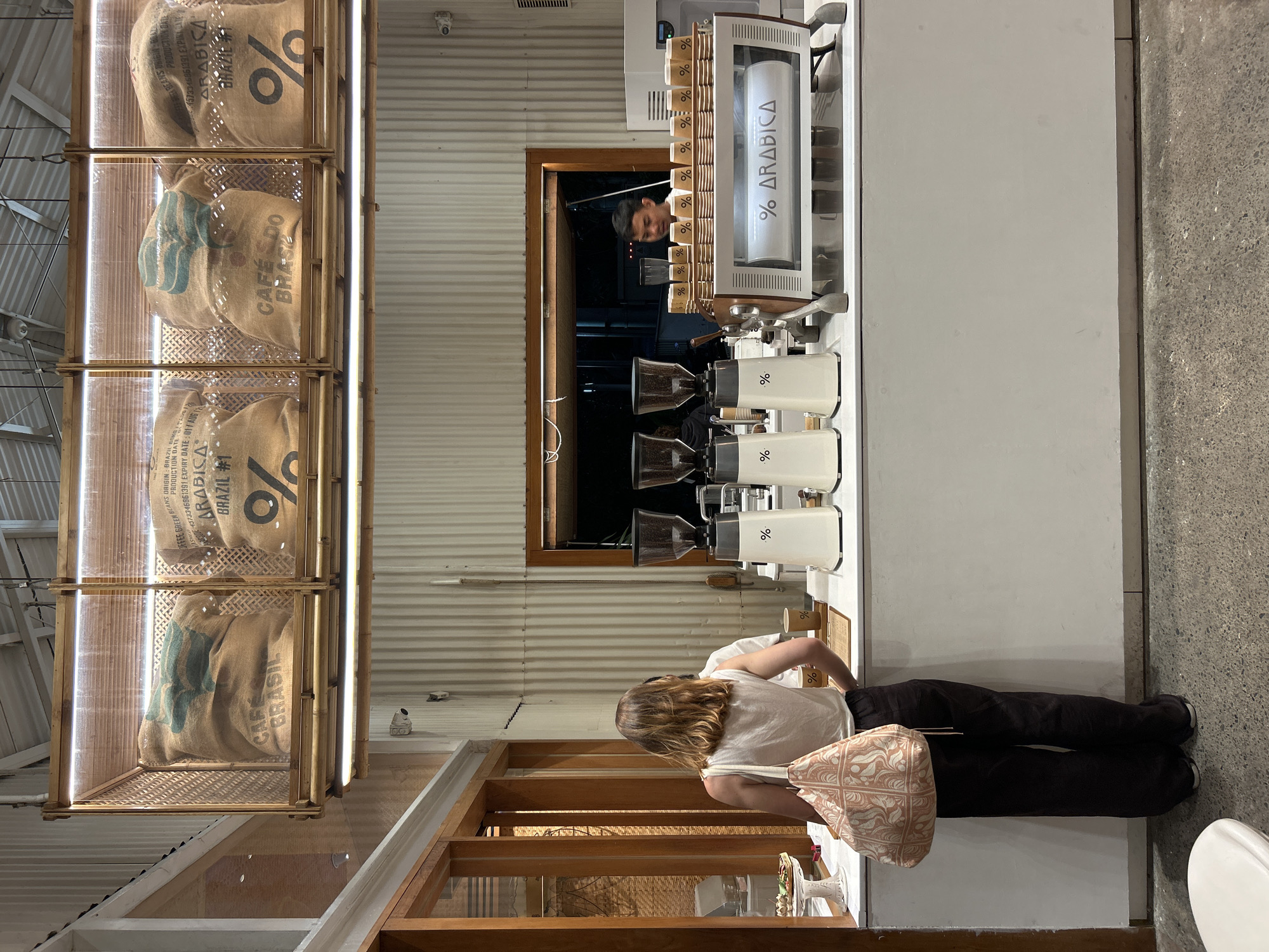 The image shows the interior of a modern cafe in Kuta Selatan, Indonesia. In the foreground, a woman with blonde hair and a white tank top stands with her back to the camera, facing the counter. She wears dark pants and carries a patterned tote bag over her left shoulder. To her right, a male barista with short dark hair, wearing a white shirt, is visible behind the counter, looking towards the woman. The counter displays multiple coffee grinders, a coffee machine with "% ARABICA" written on it, and a stack of paper cups. Above the counter, several large burlap sacks of coffee beans with the "%" logo and text like "CAFE DO BRASIL" and "ARABICA BRAZIL #1" are displayed in wooden and glass cases, illuminated by overhead lights. The cafe features corrugated metal walls and a light-colored concrete floor.