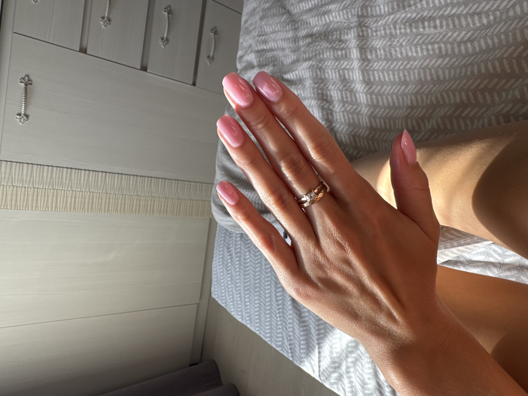 The image shows a close-up of a woman's left hand, adorned with light pink nail polish and two rings on her ring finger. One ring appears to be a band with small stones, possibly diamonds, and the other is a simple gold band. The hand is resting on a light grey and white patterned fabric, likely a duvet or bedspread. To the left of the hand, a portion of a white dresser with decorative silver handles is visible. Sunlight casts shadows and highlights across the hand and the fabric, suggesting it is daytime. The scene is indoors, likely a bedroom. The overall impression is one of personal grooming and relaxation.