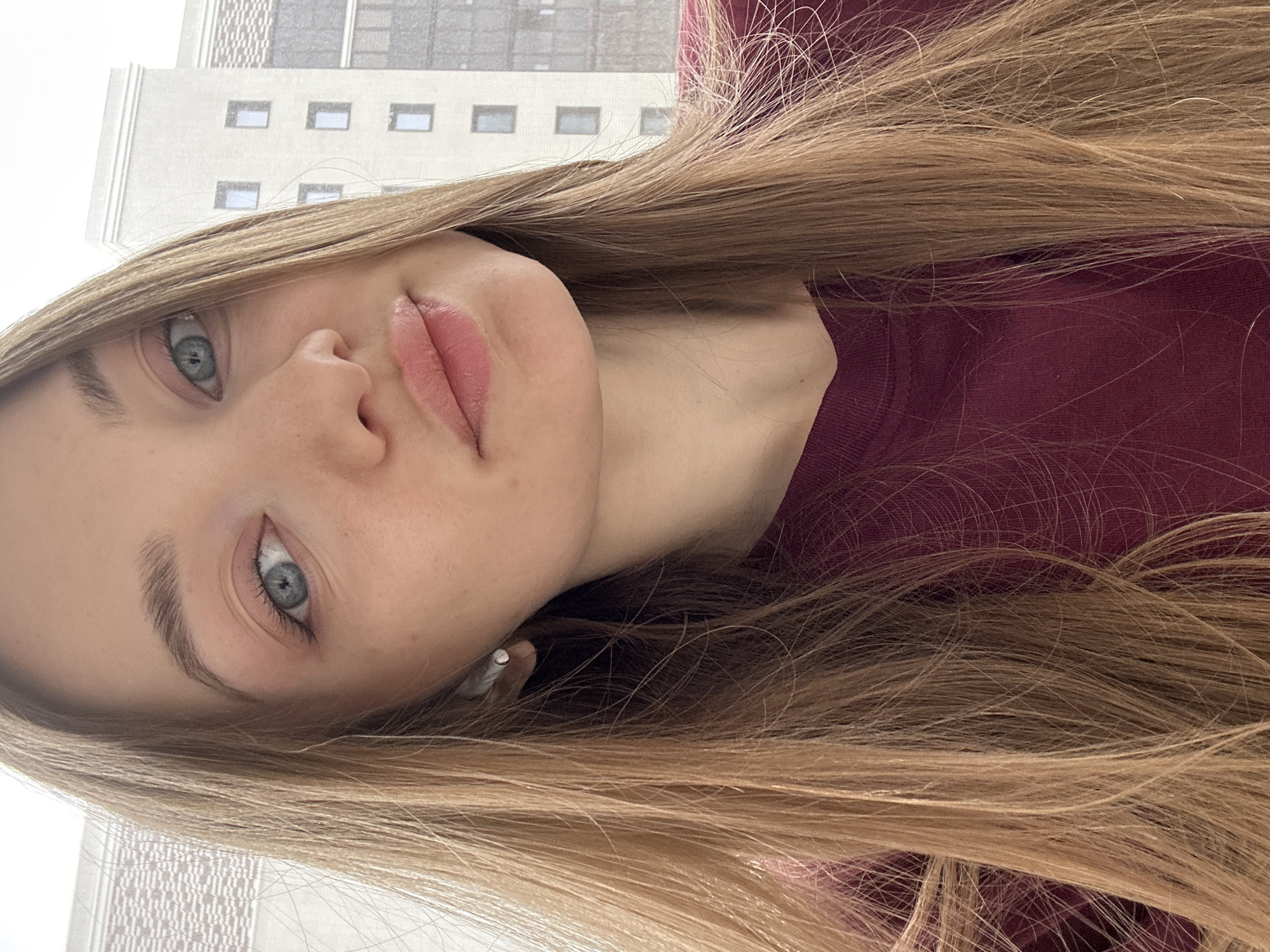 A close-up, slightly rotated portrait of a young woman with fair skin, blue eyes, and blonde hair. She is wearing a dark red or maroon top. Her expression is neutral, and she is looking directly at the camera. In the background, a building is visible, appearing to be a modern apartment complex with multiple windows, suggesting an urban environment. It is impossible to independently verify the location as Kazan, Russia from the provided image.