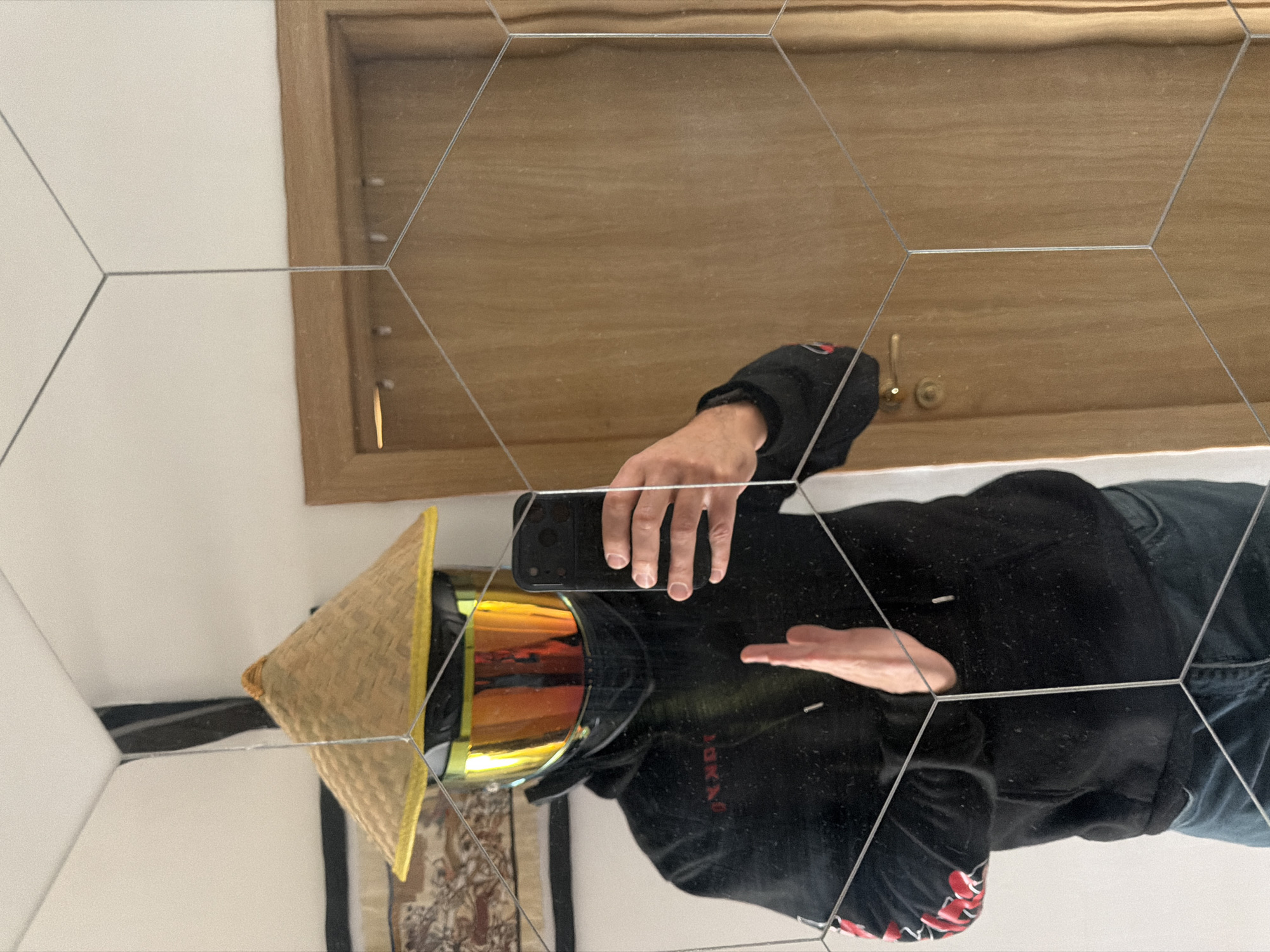 A person reflected in a multi-faceted hexagonal mirror is taking a selfie. The person is wearing a conical Asian hat and goggles with a golden-yellow lens. They are dressed in a black hoodie and jeans. The background suggests an interior setting with a wooden door, white walls, and a tapestry or rug partially visible on the left side of the frame. The location is unknown, and there is no information indicating the city of Sofia, Bulgaria.