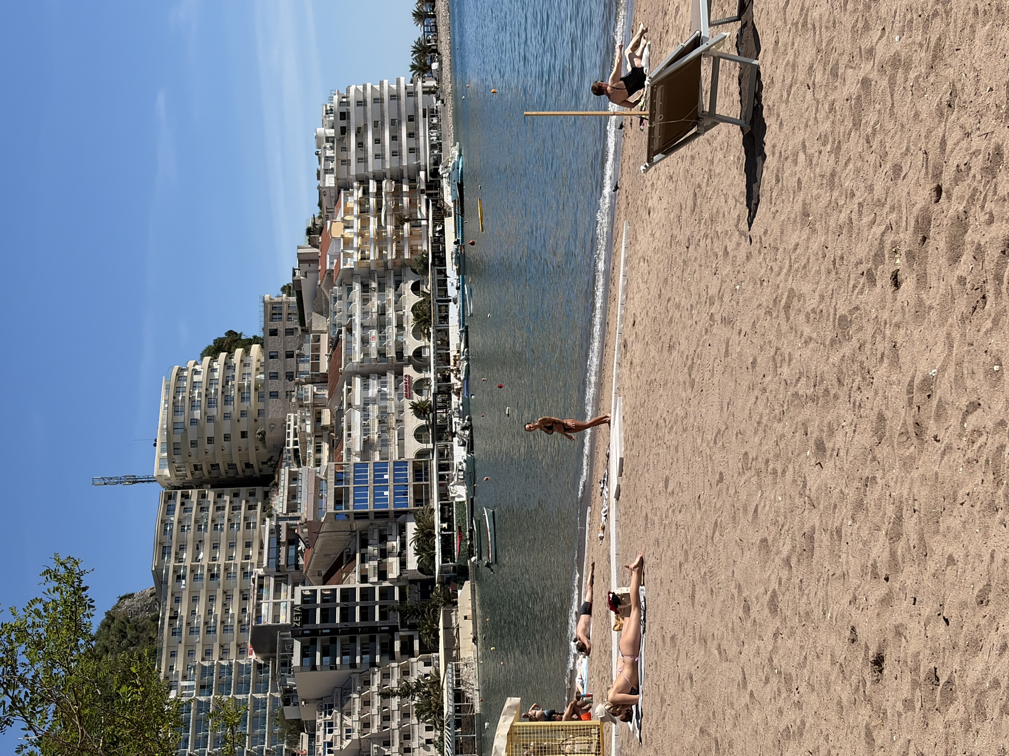 The image depicts a beach scene in Budva, Montenegro. Several people are sunbathing on the sandy beach. A woman stands near the water's edge, while others are lying on towels and beach chairs. In the background, there are tall buildings, some with solar panels, lining the coast along the Adriatic Sea. A few boats are visible in the water. The sky is clear and blue, indicating a sunny day.