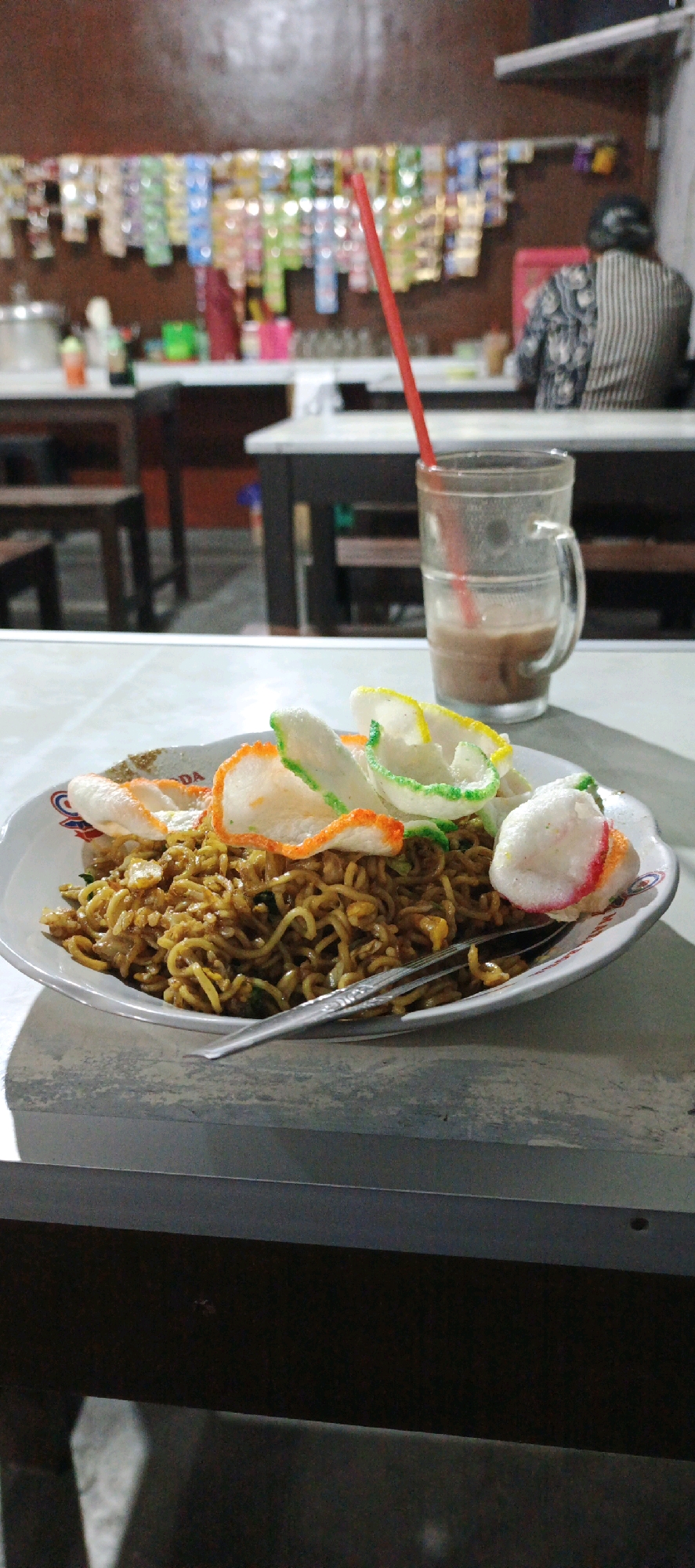 The image shows a close-up view of a plate of fried noodles, likely mie goreng, served at a roadside food stall in Banyuraden, Indonesia. The noodles are topped with colorful crackers, some with orange and green accents. To the right of the plate, a glass of iced coffee or chocolate milk with a red straw is visible. In the background, shelves are stocked with various packaged snacks and drinks, a common sight at Indonesian street food establishments. A person wearing a patterned shirt is partially visible in the far right background, suggesting a busy eatery. The lighting is dim, indicating it is likely evening or night. The setting appears to be an open-air or semi-enclosed stall, typical of Indonesian street food culture.