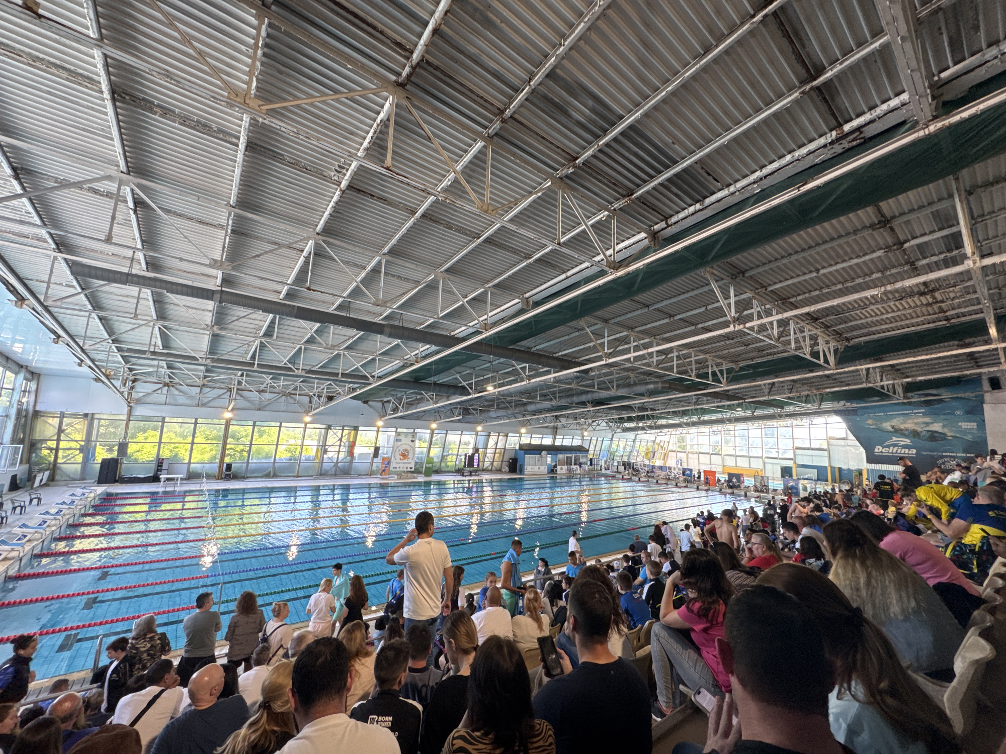 The image depicts the interior of a swimming pool complex, likely in Sofia, Bulgaria. A competition is underway in the pool, with multiple lanes visible and swimmers in the water. Spectators fill the stands, observing the event. The building has a large, open roof structure with metal supports and corrugated roofing material. Large windows line one side of the pool, providing natural light. A mural or sign with the word "Delfina" is visible in the background. The people in the stands are of varied ages and are dressed casually. Some are taking pictures or videos of the event.