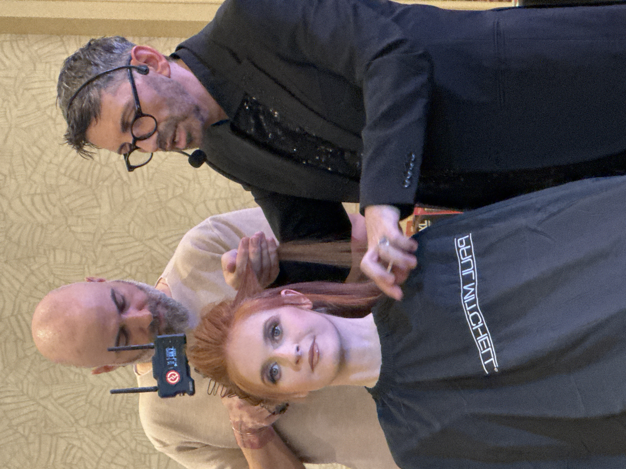 The image shows a woman with red hair, wearing a black cape with white text, receiving a haircut. One man in a black suit and glasses is cutting her hair, while another man in a tan shirt is holding the hair. The location is Unknown, Unknown.