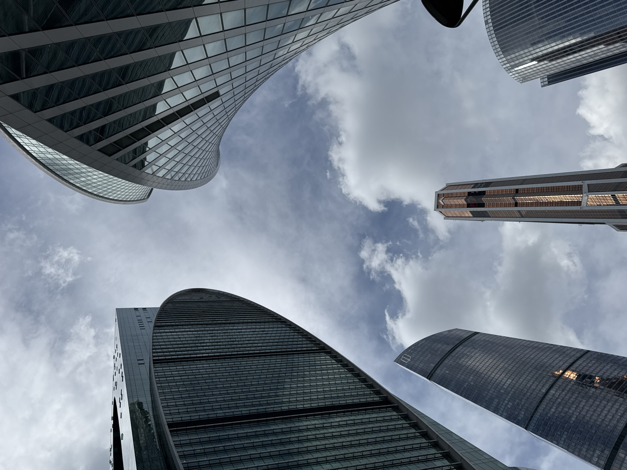 The image is a low-angle perspective of several skyscrapers against a cloudy sky. The skyscrapers feature modern architectural designs with glass facades. The sky is predominantly gray with patches of light blue visible between the clouds. There are no people visible in the frame. While the image depicts modern skyscrapers, there is no information present to determine that the location is in the city of Khimki, Russia.
