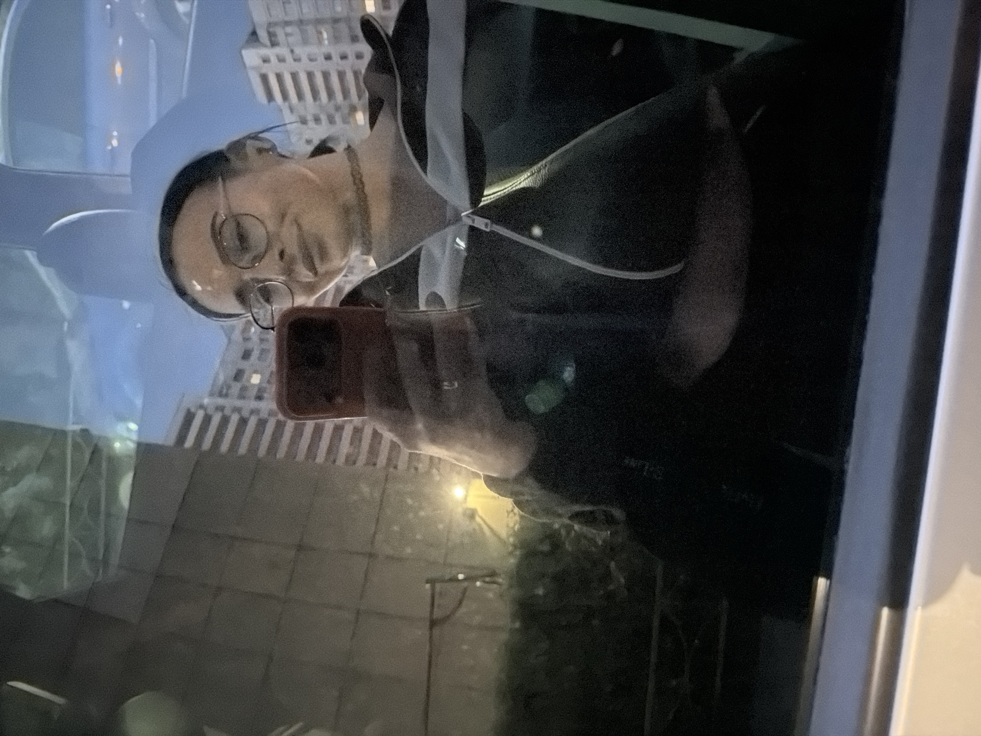 The image captures a reflection of a person taking a photo with a smartphone, set against an urban nightscape in Saint Petersburg, Russia. The individual, appearing to be female, is positioned centrally, wearing round eyeglasses and a dark jacket or hoodie with a distinct white vertical stripe. They are holding a smartphone with a reddish-brown case in their right hand, angled towards themselves, suggesting a selfie or a reflective shot. A ring is visible on their ring finger. Their expression is neutral, looking directly ahead into the reflective surface.
The background, visible through the reflection, depicts a city environment at night or dusk, with illuminated multi-story buildings, possibly residential or commercial structures, lining the horizon. A street light or other outdoor light source casts a bright yellow glow in the lower part of the scene. The overall lighting is dim, characteristic of evening, with artificial lights providing illumination. A faint, indecipherable white text or logo, possibly "S-Lab," is visible on the person's left sleeve (right in the reflection). The image was likely taken indoors looking out through a window, or against a large reflective surface.