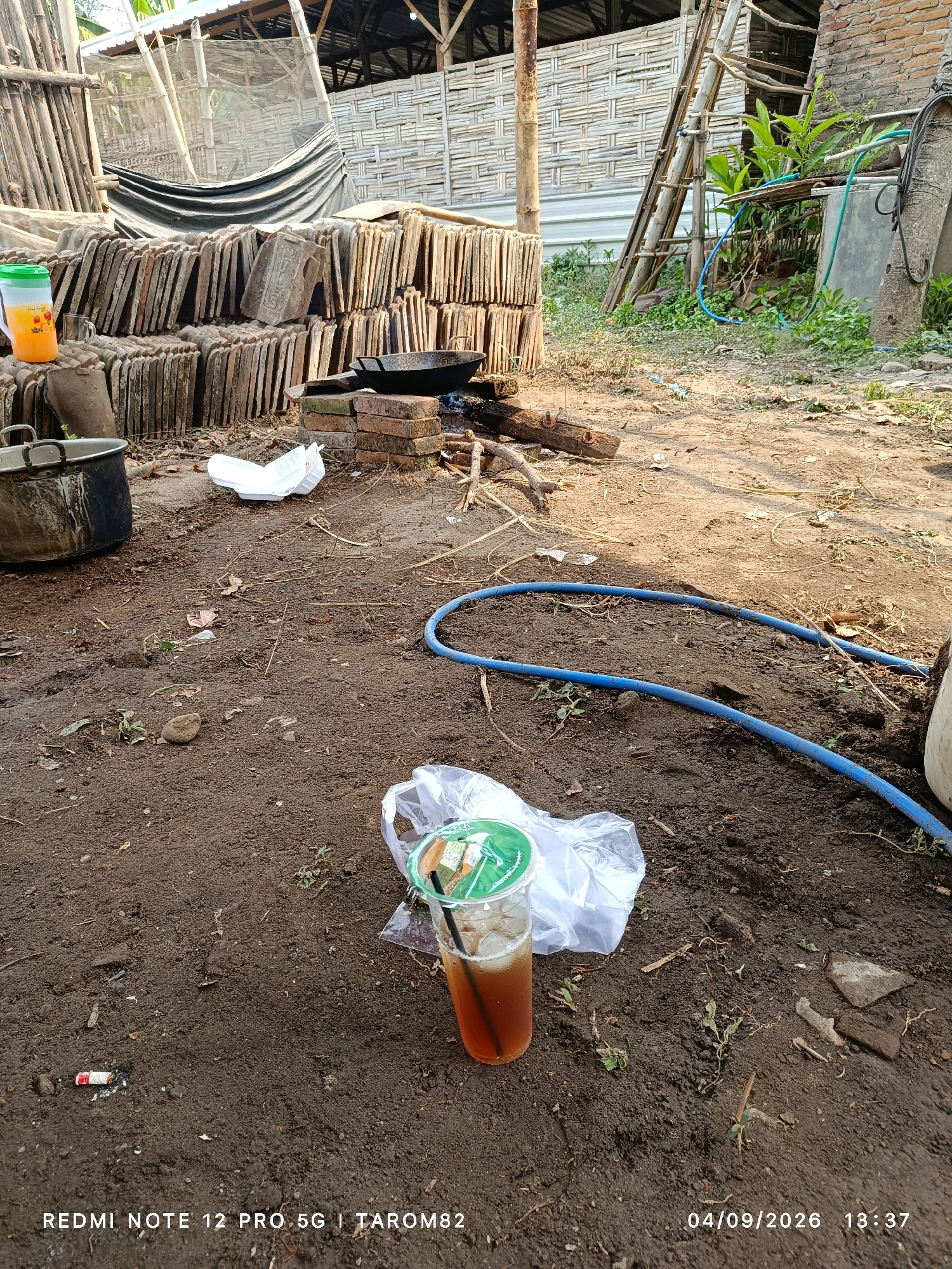 The image captures an outdoor scene in what appears to be a rural or semi-rural setting, possibly a yard or workshop area, with the timestamp and text overlay indicating the location is in Rejoagung, Indonesia, on 04/09/2026 at 13:37. The ground is predominantly dirt with scattered debris, including dry leaves, twigs, and some small rocks. In the foreground, a plastic cup with a brown beverage and ice, accompanied by a straw, sits on the ground next to a white plastic bag. A used cigarette butt with a red tip lies a short distance to the left of the cup.
In the mid-ground, a blue hose is visible coiled and extending across the dirt. To the left, a large black pot or cauldron rests on the ground. Further back, stacks of roof tiles are arranged vertically, forming a substantial barrier. A black frying pan is placed on a makeshift stove constructed from bricks, with embers visible underneath. A smaller glass or cup is placed near the stacks of tiles, next to a plastic jug filled with an orange liquid.
The background features a structure with a thatched or woven roof supported by bamboo poles. A ladder leans against this structure, and lush green vegetation is visible growing around it. Part of a tree trunk is also present on the right side of the image. The overall impression is one of a working area or a place where cooking or construction activities may occur, with a casual accumulation of items and a somewhat untidy environment.