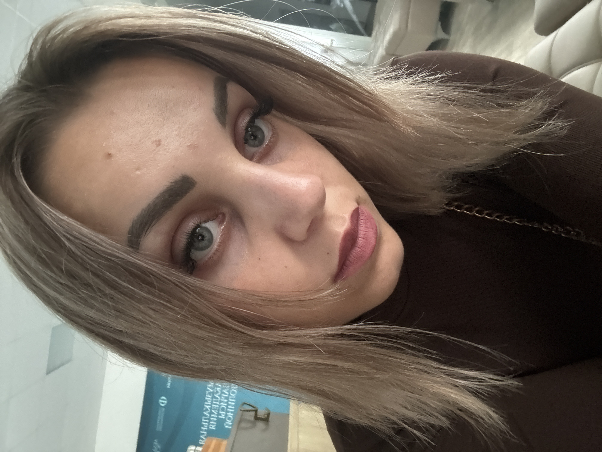 A young woman with light brown hair and light blue eyes is captured in a close-up, slightly downward-angled selfie. She is wearing a dark brown turtleneck shirt with a subtle gold chain visible over her shoulder. Her makeup includes defined eyebrows, eyeliner, mascara, and a matte pink lipstick. Her skin appears fair with a few small blemishes on her forehead. The background is blurred, but a large, light blue banner with Cyrillic text and a circular logo is visible on the left side. The overall lighting suggests an indoor setting, possibly an office or public space. The text on the banner is partially obscured but includes what appears to be "РАННЕЛЙ". This banner is consistent with those displayed at the Sakhalin Regional Universal Scientific Library in Yuzhno-Sakhalinsk, Russia, which often hosts events and exhibitions, suggesting the location. The woman is the sole subject and focus of the image.