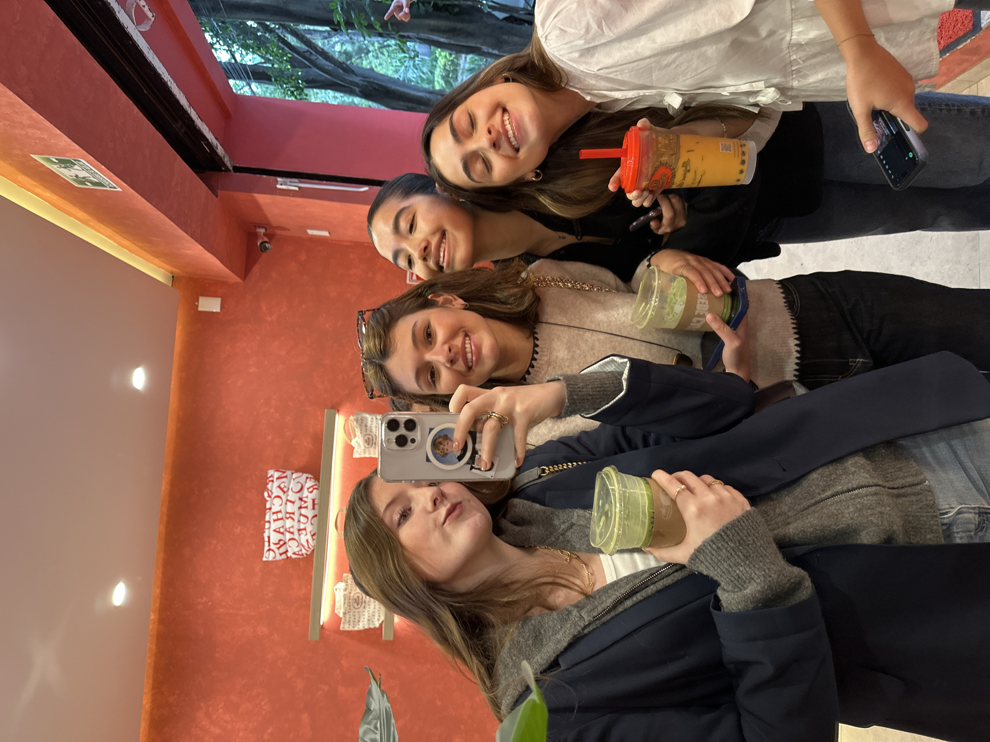 Four young women are posing for a selfie indoors. The woman in the foreground is holding a smartphone and a green beverage. The other three women are smiling and holding beverages. The background includes a red wall with light fixtures and bags on display. Tree branches are visible through a window above.