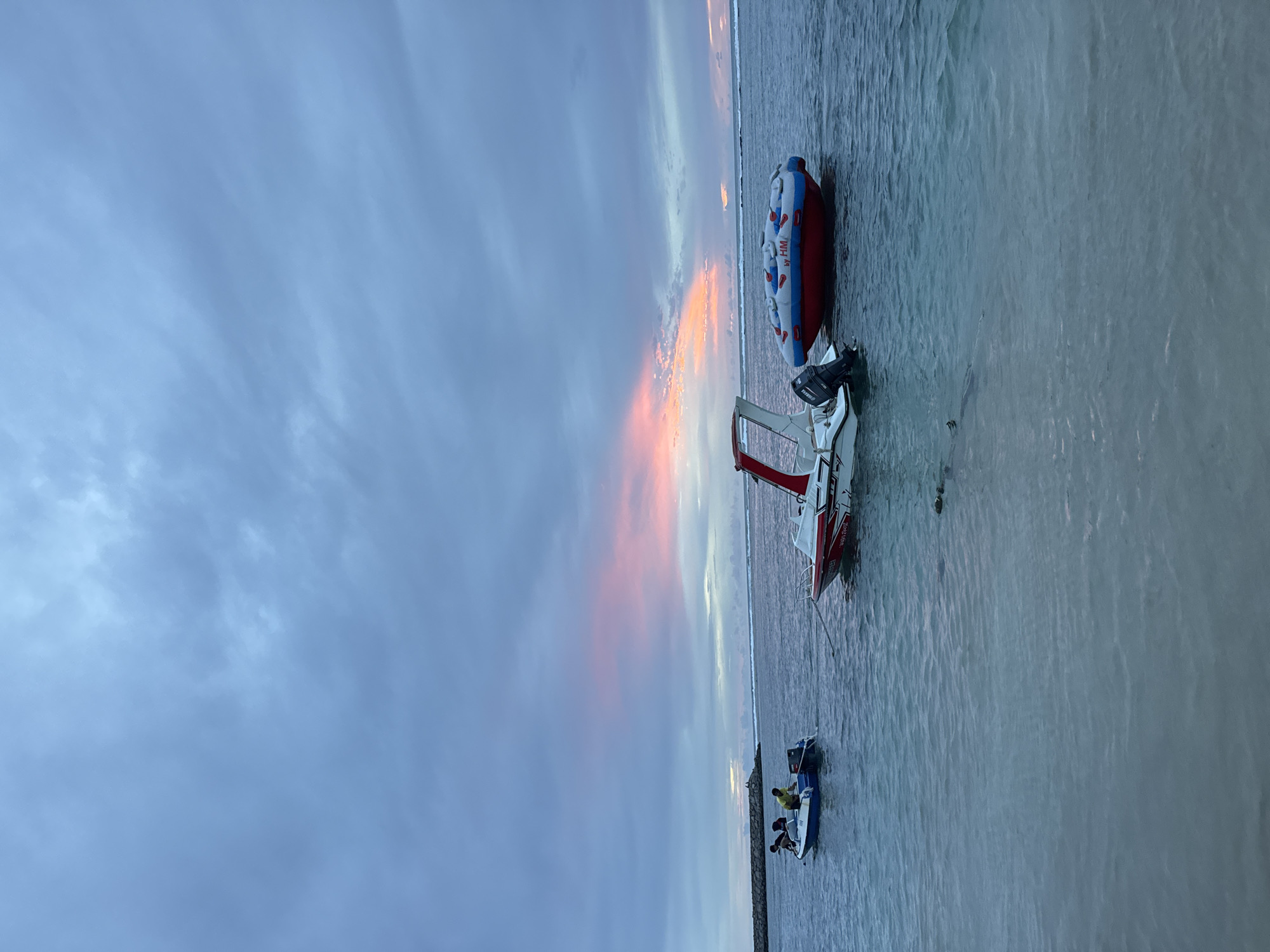 The image shows a serene seascape at dusk, likely in Meunasahbale, Indonesia, with a hint of pink and orange hues in the clouds above. In the foreground, the calm, clear turquoise water laps gently against a sandy shore. Three boats are anchored in the shallow water. To the left, a small white and blue motorboat carries three individuals, one of whom appears to be wearing a yellow shirt. A second, slightly larger white motorboat with red accents is positioned in the middle of the frame, its outboard motor visible. To the right, a large red and blue inflatable tube, possibly for water sports, floats on the surface. In the distance, a rocky breakwater extends into the sea, with a faint outline of land or the horizon visible beyond. The sky is predominantly overcast with a blend of gray and muted blue, illuminated by the soft glow of the setting sun.