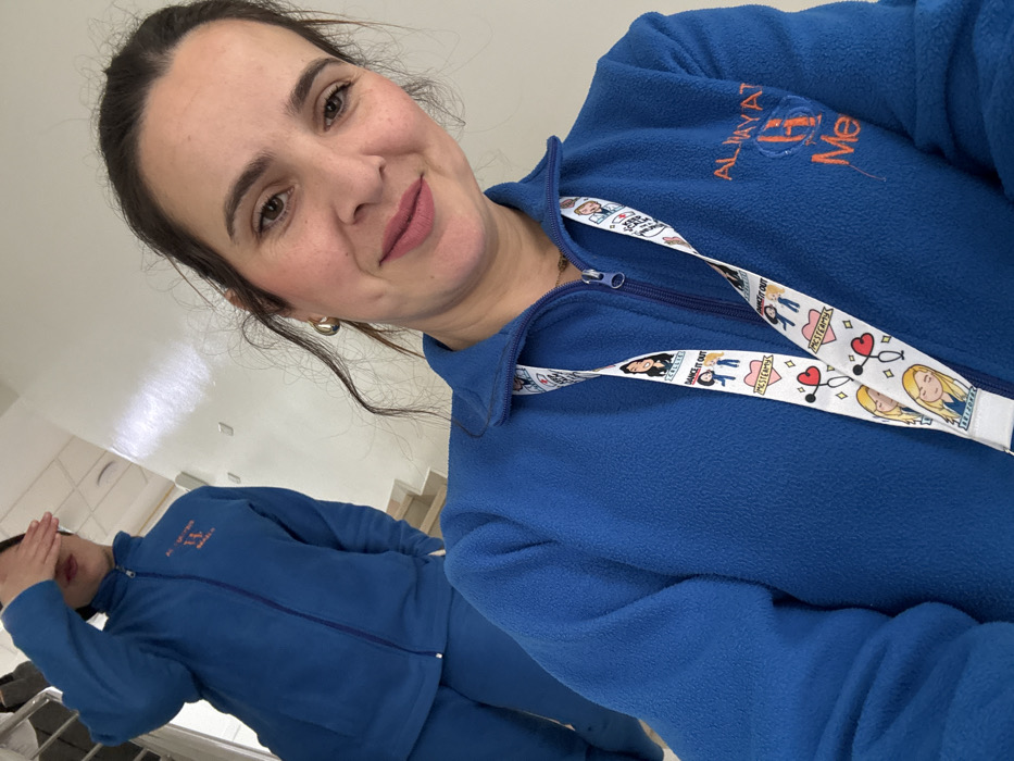 The image captures two women indoors, likely within a medical facility in Meknes, Morocco. The foreground subject, a woman with dark hair, a faint smile, and light makeup, is taking a selfie. She wears a blue fleece zip-up jacket with an orange embroidered logo on the right chest area that reads "AL HAYAT H Me," where the 'H' is stylized as a medical cross, identifying the location as a healthcare institution named "Al Hayat" in Meknes. A white lanyard adorned with cartoon figures, medical symbols (stethoscopes, hearts), and text such as "DANCE IT OUT" and "MESTEAM" hangs around her neck. In the background, a second woman, similarly dressed in a blue fleece zip-up jacket and matching blue trousers, stands with her right hand covering her eyes, obscuring her face. The environment features plain white walls and a visible staircase, indicating an interior space with multiple levels.