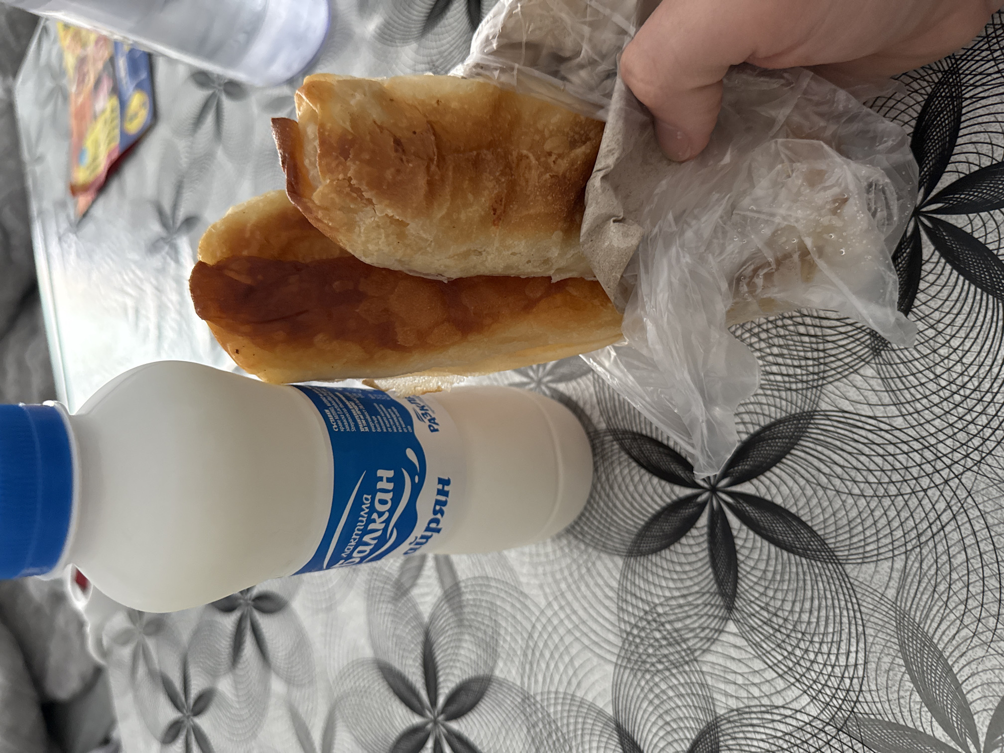 The image shows a snack of pastries in a plastic bag and a bottle of Bulgarian dairy drink "Ayran" produced by Laktima Balkan. The background is a table with a floral pattern. While Stara Zagora is mentioned in the Laktima Balkan’s address, it is not clear from the image that this image was taken in that city.