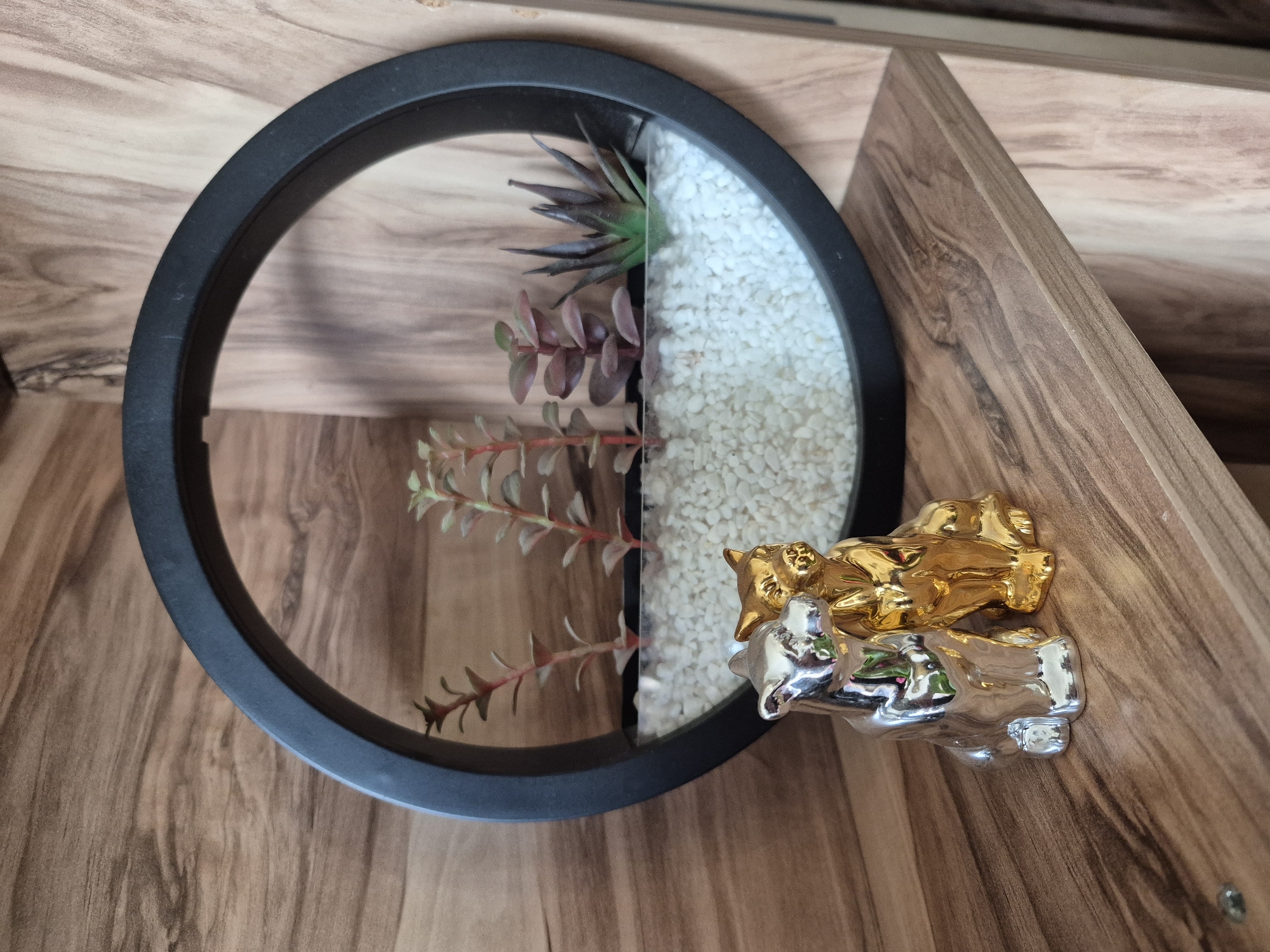 A top-down shot depicts a circular, black-rimmed, decorative piece containing artificial plants and white stones, bisected vertically. To the right are two metal figures of embracing individuals, one gold and one silver, resting on a wooden surface. The wooden surface features a linear grain pattern. The location of the scene is not discernible from the visual information provided.