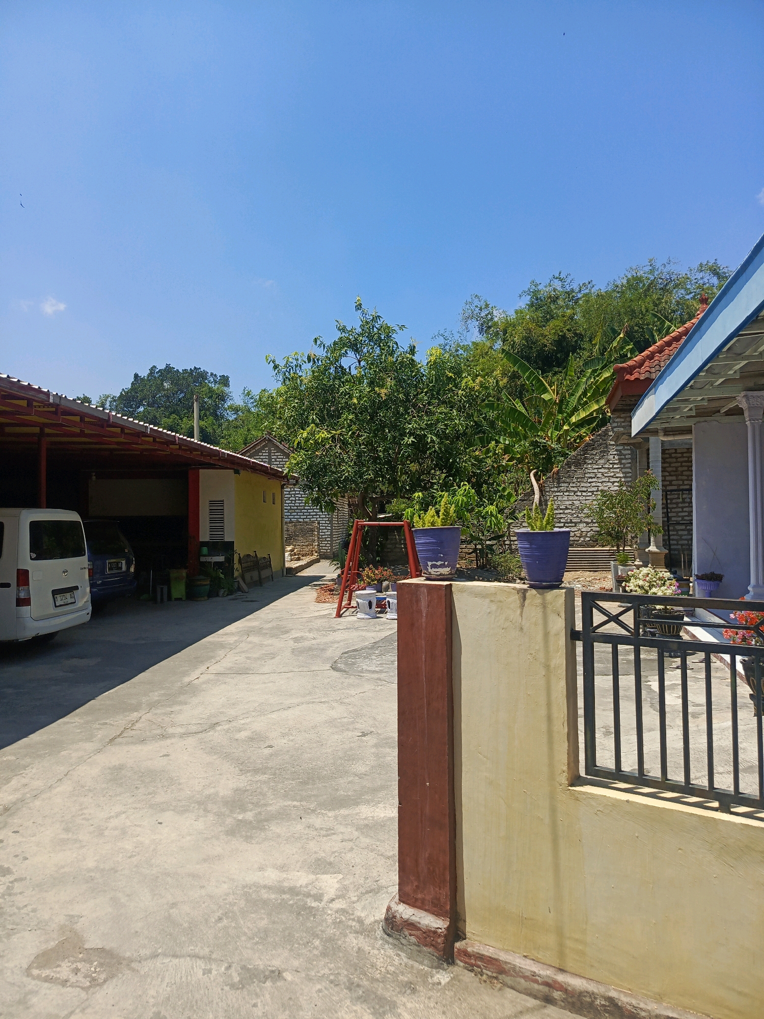The image depicts a residential scene in Pamekasan, Indonesia, under a clear blue sky with scattered birds. In the foreground, a light-colored concrete courtyard is visible, marked with cracks and a small puddle. To the right, a portion of a building with a light yellow wall and a dark brown trim is shown, featuring a black metal fence with vertical bars. Behind this, a covered patio area shelters a white van and a blue vehicle. Further into the scene, a yellow building with a red-trimmed roof is situated, and beyond that, a wall constructed of rough-hewn stones. A red swing set with two seats is positioned in the middle ground. Several potted plants, including two large blue pots with spiky green plants and smaller pots with flowering plants, are placed around the courtyard. Lush green trees and vegetation dominate the background, with a more distant hillside covered in trees. The overall impression is one of a rural or semi-rural residential setting.