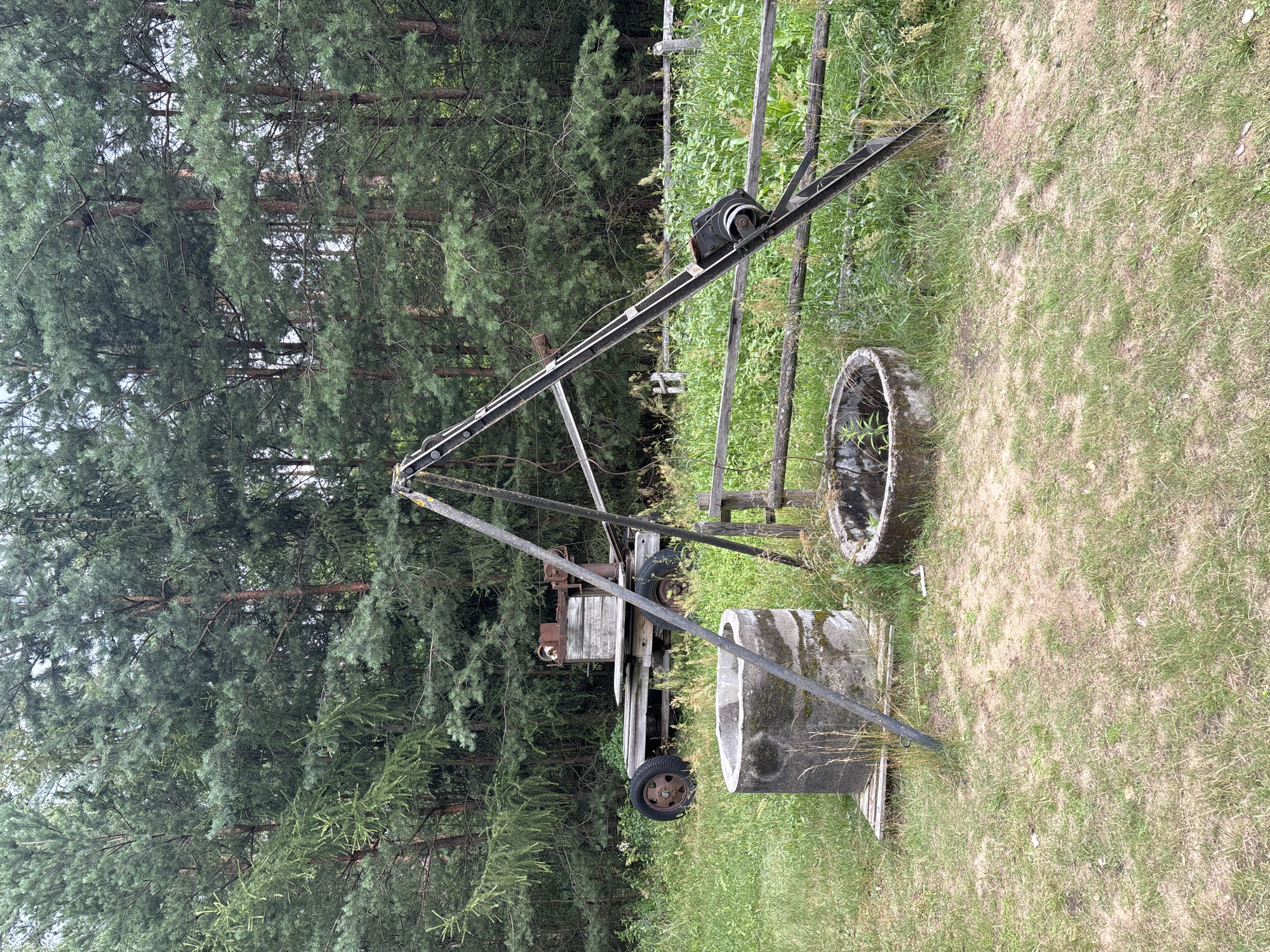 An outdoor scene in Sielachowskie, Poland, features a grassy clearing with a backdrop of dense evergreen trees. In the foreground, two circular concrete structures, likely old wells, are visible. To the left is a larger, taller well, with a wooden pallet beneath it. To the right is a smaller, shallower well. A rustic wooden fence runs horizontally across the mid-ground. A triangular metal structure, possibly a makeshift crane or derrick, stands prominently in the center, with a pulley mechanism attached to its upper section. Behind the wells and fence, a weathered wooden trailer is parked. It has two large wheels, one of which is partially obscured. On the trailer, a piece of rusty machinery with a wooden component is mounted. The overall impression is one of an abandoned or disused area, likely on the outskirts of the settlement.