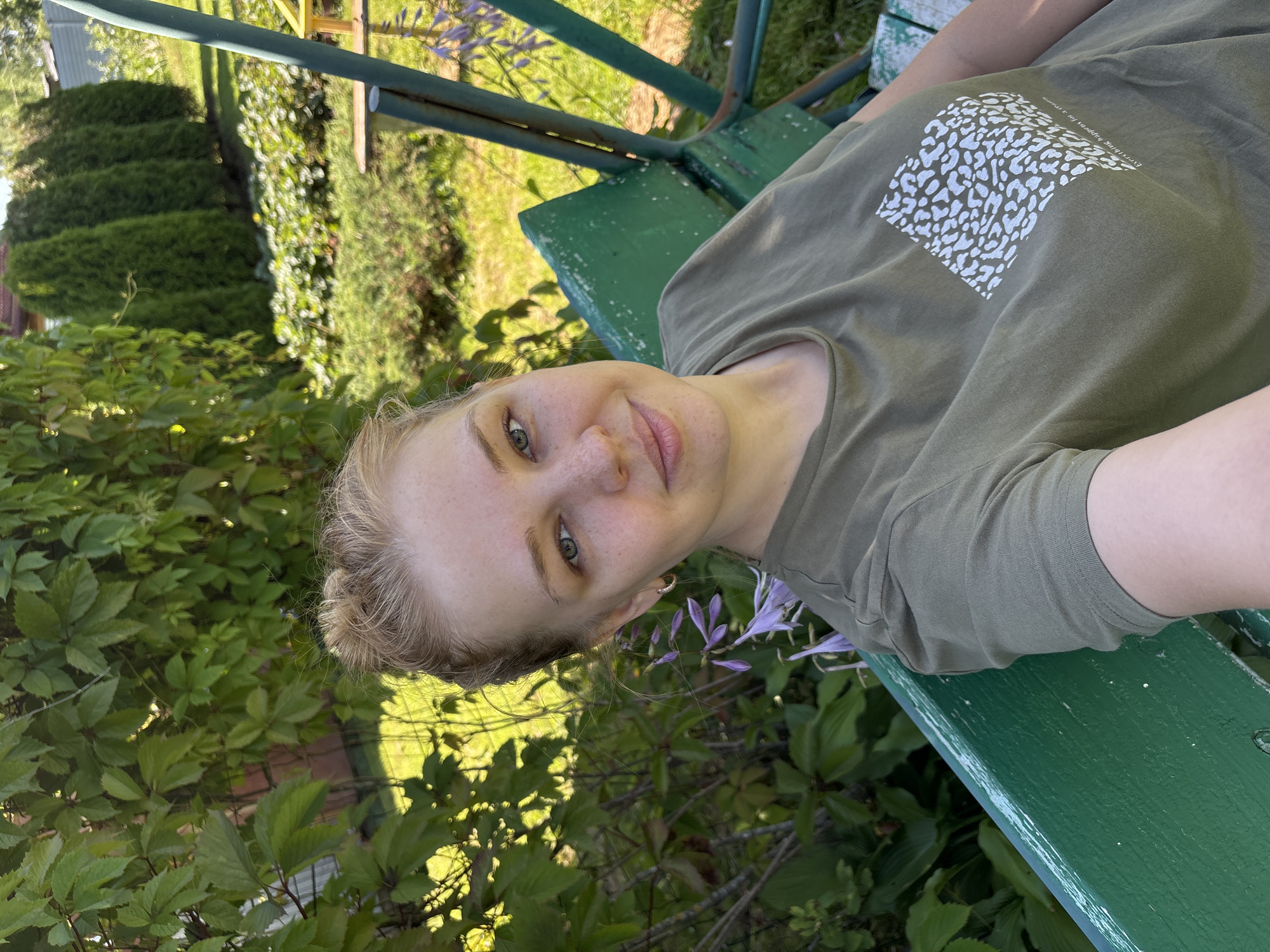 A young woman with blonde hair pulled up in a bun is sitting on a green swing set in a garden. She is wearing a green t-shirt with a white, leopard-print-like pattern on the front. The swing set is old and weathered, with chipped green paint. Behind the woman, there are lush green trees and bushes, a fence, and a glimpse of a house. In the background, several tall, slender evergreen trees are visible. The scene appears to be set in a rural area, possibly the village of Dunilovo, Russia.