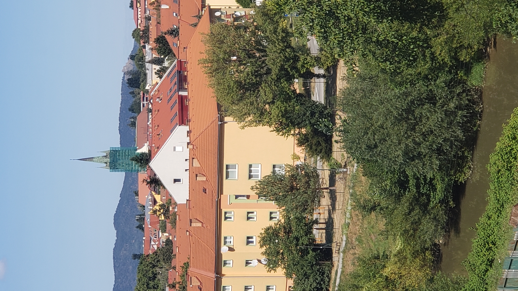 This is a vertical, medium-angle, eye-level, full shot of a residential area in the city of Prešov, Slovakia, on a clear, sunny day. In the foreground, a body of water, likely a river, is visible, bordered by lush green trees and bushes. Beyond the water, a grassy area with scattered trees and benches is seen, leading up to a yellow apartment building with numerous windows and satellite dishes. Behind the apartment building, several other buildings with orange tiled roofs are present, interspersed with trees. In the distance, a tall, slender church tower with a green spire is partially covered by scaffolding. Further in the background, a range of blue-hued mountains forms the horizon under a clear blue sky. No people are visible in the frame.