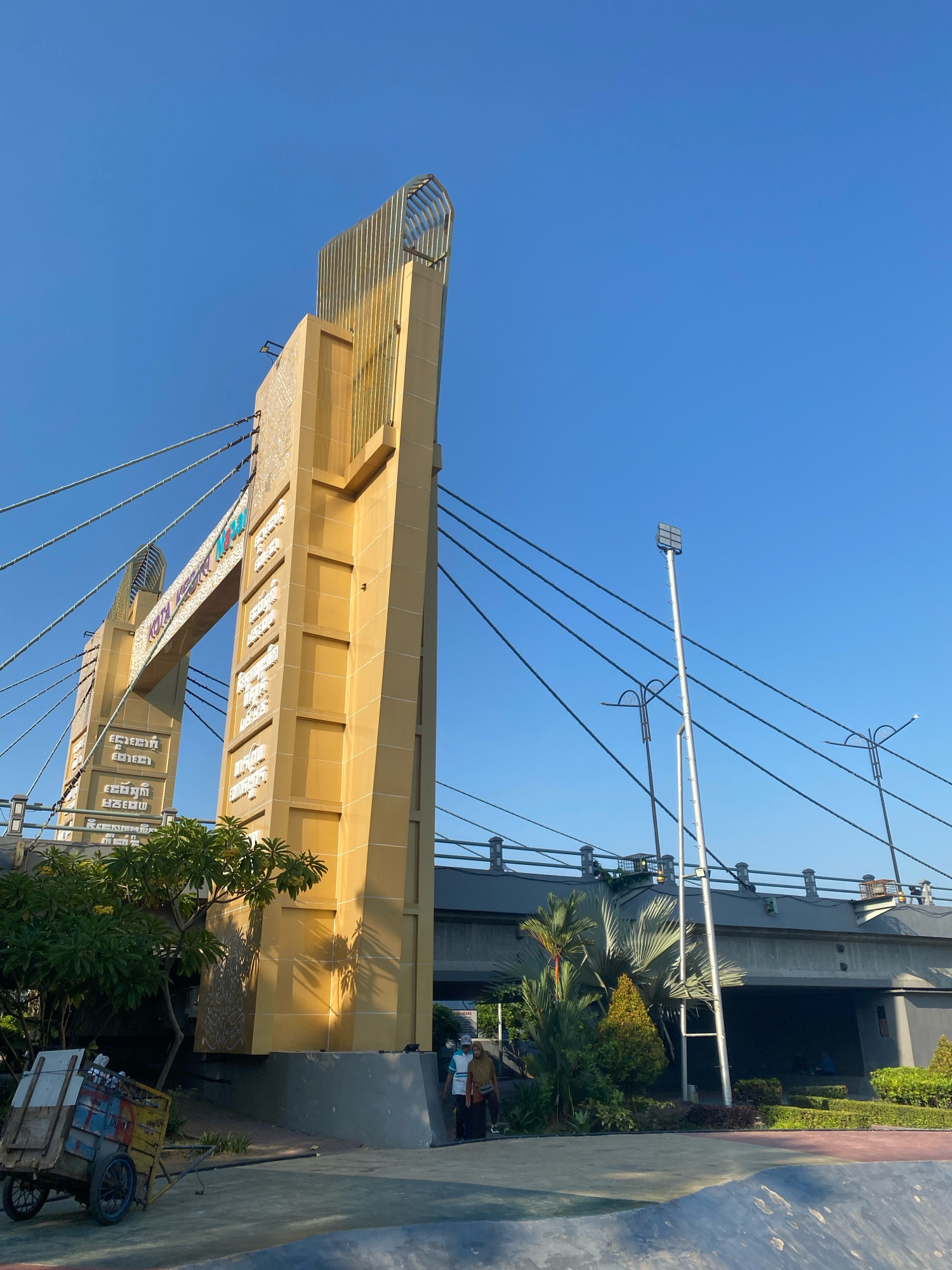 The image shows a view of the Jembatan Brawijaya (Brawijaya Bridge) in Kediri City, Indonesia. The bridge features distinctive golden towers with intricate patterns and is suspended by cables against a clear blue sky. In the foreground, a colorful, wheeled cart is parked near some greenery. To the right, a multi-level bridge structure is visible, with streetlights and lush vegetation. Two people, one wearing a light blue shirt and the other a brown hijab, are walking near the base of the golden tower. The overall scene depicts a sunny day with architectural elements of the city and its surrounding environment.