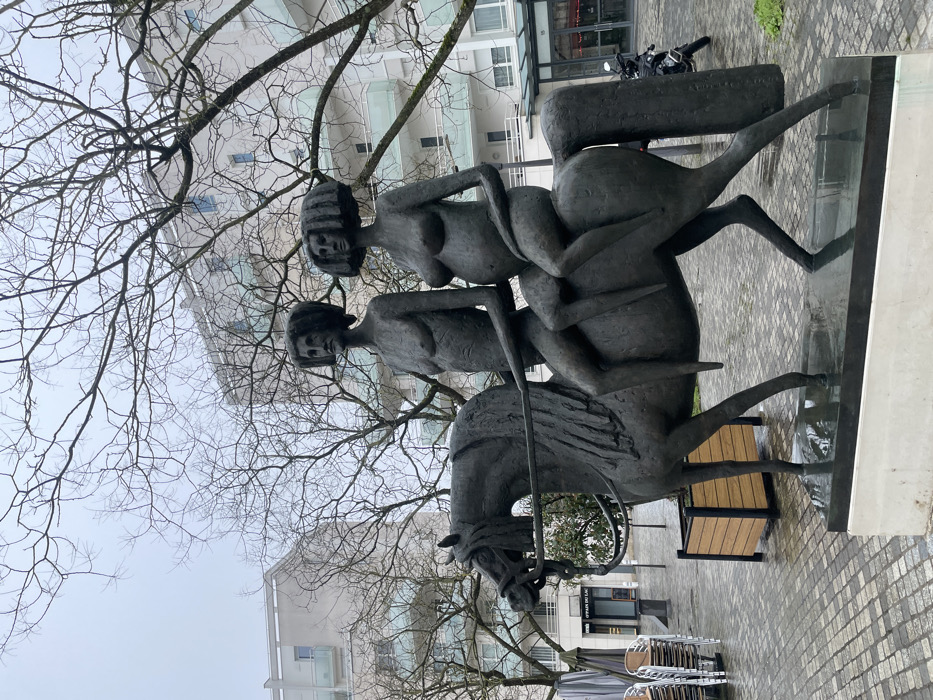 A large bronze sculpture, "Les Cavaliers du Lac," is situated in an urban square in Montigny-le-Bretonneux, France. The sculpture depicts a horse carrying two nude female figures. The front figure, a woman with prominent breasts and stylized, broad hair, rides astride, gripping reins. The rear figure, also a nude woman with similar stylized hair, is visibly pregnant and seated behind the first rider. Both figures exhibit distinct, stylized facial features. The horse is sculpted with a bridle, mane, and a long, solid tail. The sculpture rests on a light-colored rectangular plinth. The ground consists of wet, cobblestone-patterned paving. In the background, multi-story modern buildings with light-colored facades and balconies are visible. Numerous deciduous trees with bare branches are interspersed throughout the scene, indicative of a colder season. To the left, stacked outdoor café chairs and tables are present. To the right, a black motorcycle is parked near a building entrance. The sky is uniformly overcast and grey.
