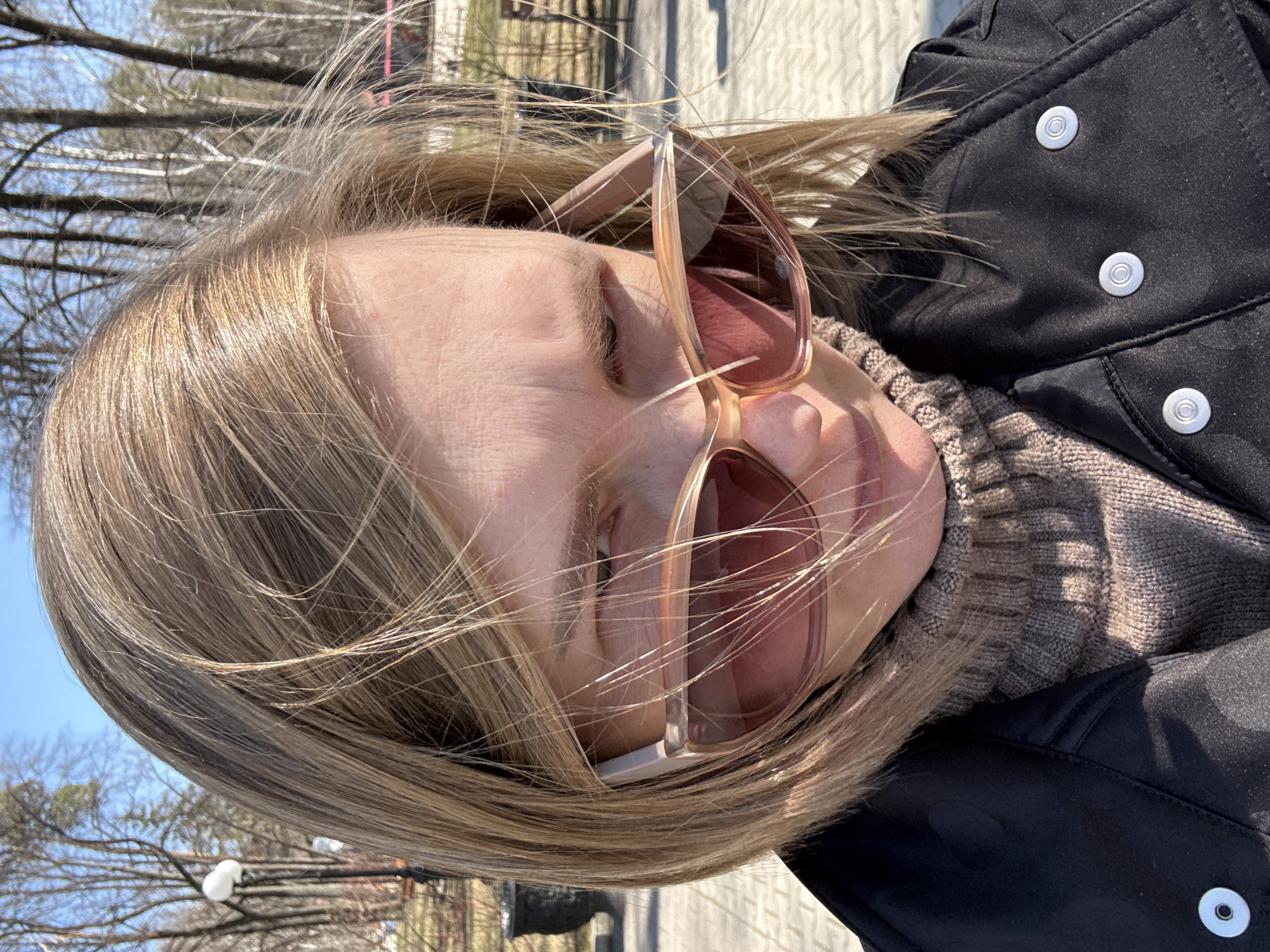 A person with light brown hair and wearing sunglasses is the primary subject of the image. They are also wearing a brown turtleneck sweater and a black coat with silver buttons. The background shows trees and what appears to be a paved area, possibly a park or walkway. It is difficult to conclusively determine the exact location in Yekaterinburg, Russia from the image alone without more contextual clues.