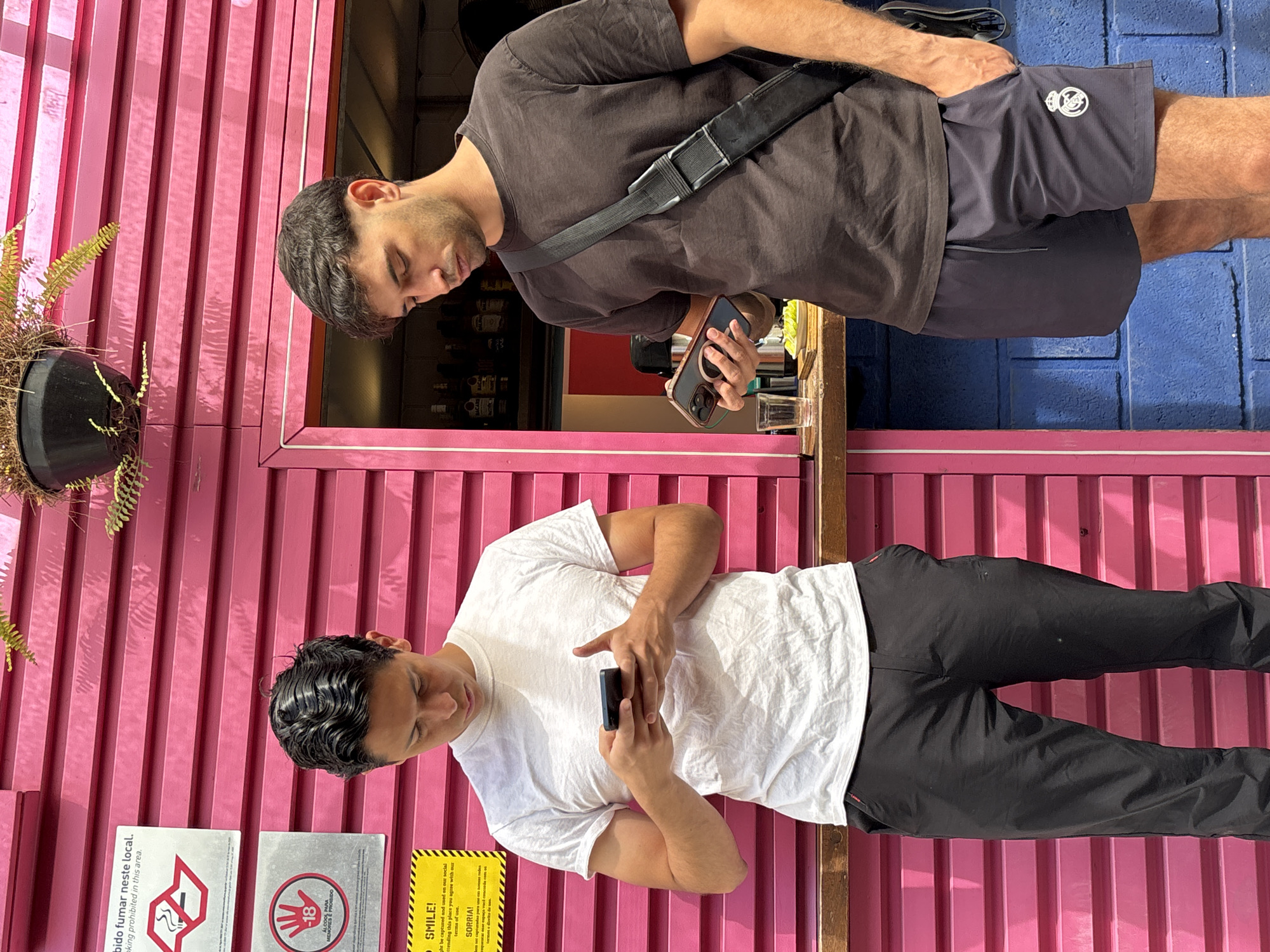 The image captures two men in front of a pink-painted corrugated metal structure. The man at the top of the image wears a dark brown shirt, gray shorts with a logo, and a black shoulder strap, looking at his phone. The second man, dressed in a white t-shirt and black pants, also focuses on his phone. On the left side of the structure, there's a hanging plant. Various signs are visible, one prohibiting smoking and another indicating that the area is under video surveillance and by entering the area, one agrees to having their image used. The image appears to have been taken outdoors, in São Paulo, Brazil.