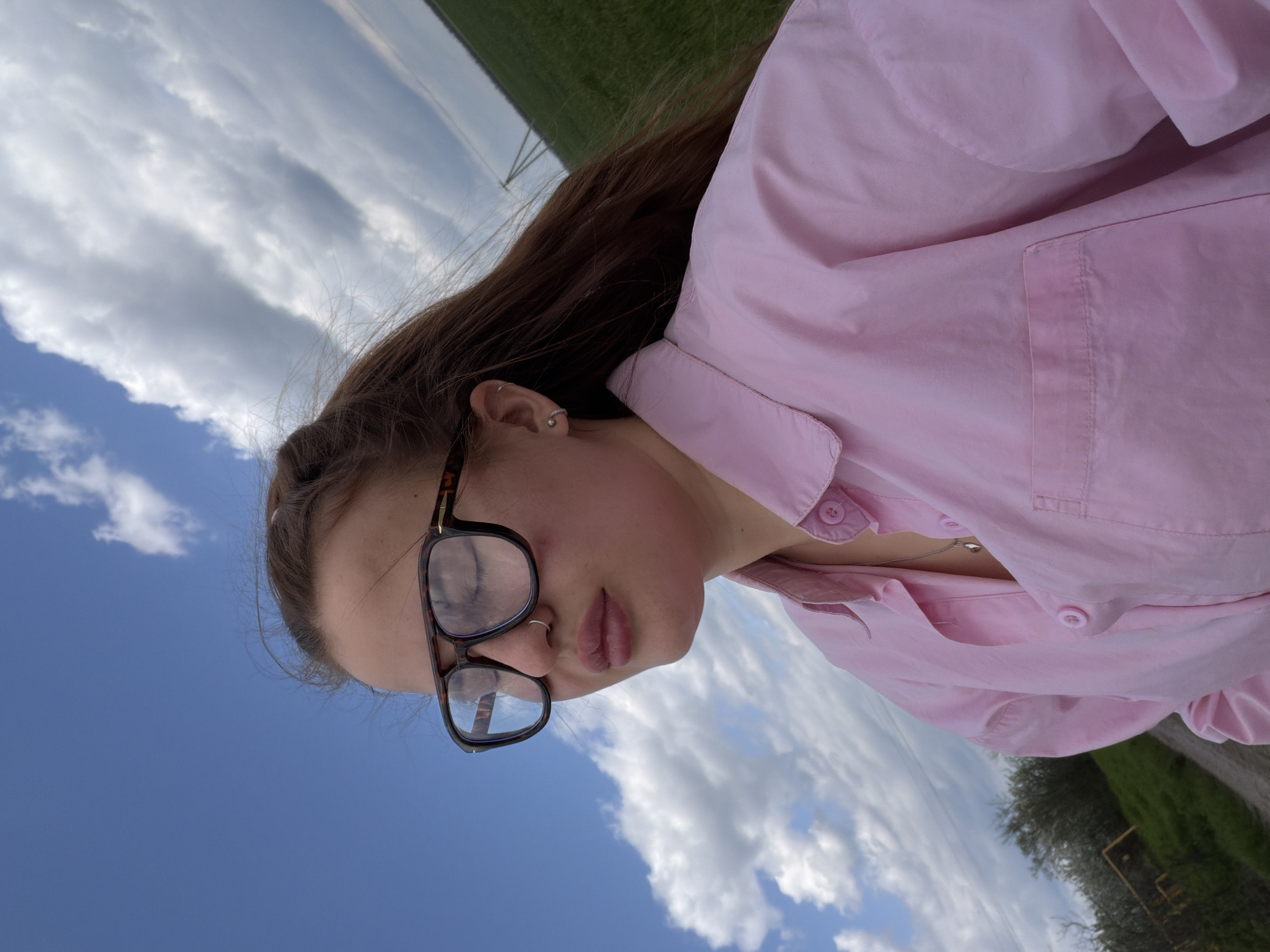 A rotated, upward-angled photo captures a young person, likely female, outdoors under a bright blue sky with scattered white cumulus clouds. The subject occupies the right and center of the frame, positioned diagonally. She has long, dark brown hair, worn down, and is looking slightly downwards with a neutral expression. She wears large, dark-rimmed glasses, a small silver hoop nose piercing, and a silver stud and hoop in her ear. Her attire consists of a light pink, long-sleeved button-up shirt, with some buttons undone to reveal a silver chain necklace.
The background features a green grassy field on the upper left side, with a faint metal structure visible in the distance. The lower right background shows dense foliage from trees and what appears to be a paved path or road. The daylight is bright, suggesting a sunny day with pleasant weather. There is no visible text. The setting appears to be a rural or suburban outdoor area in Novopokrovskaya, Russia.