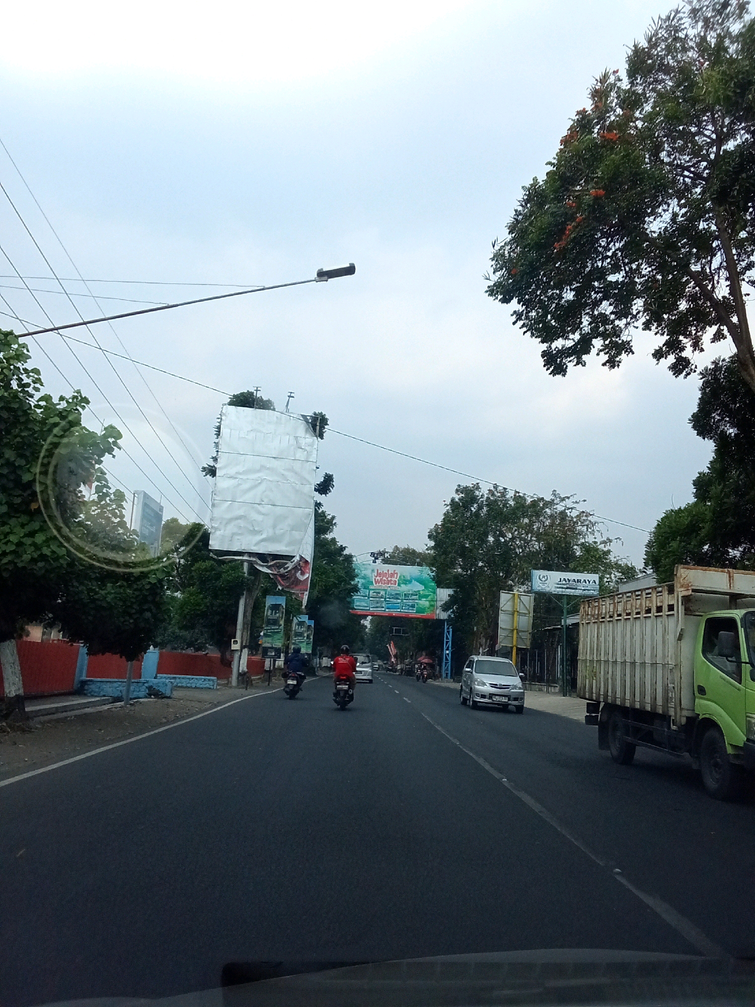 The image captures a street scene in Ngasem, Indonesia, under an overcast sky. A paved road dominates the foreground, with a white line marking the lane. Several vehicles are visible: a silver car is driving in the left lane, and a green truck is in the right lane. Two motorcycles are also present, one with a rider in red and another with a rider in dark clothing, both traveling in the left lane.
On the left side of the road, a red and blue wall borders a sidewalk. Several trees with green foliage are scattered along the roadside. A large, dilapidated billboard covered in reflective material stands on the left, and another billboard advertising "Jelajah Wisata" is visible further down on the left. On the right, a billboard with the text "JAYARAYA" is mounted on a pole. Power lines crisscross the sky, and a street lamp extends from the left side of the frame. The overall environment suggests a typical urban or suburban setting in Indonesia.