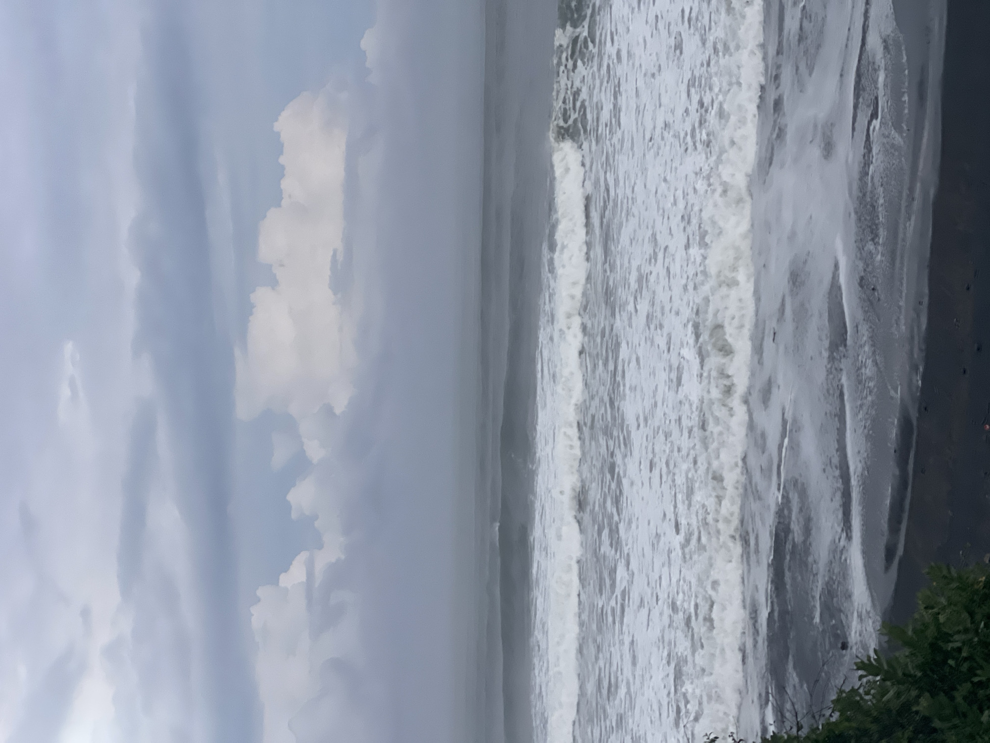 The image shows a cloudy sky over a dark sand beach with moderate waves crashing onto the shore. In the foreground, dark green foliage is visible on the left side of the frame. The ocean stretches to the horizon, with several layers of waves visible, the closest ones breaking and forming white foam. The sky is overcast with large, diffuse clouds, some of which appear white and illuminated, while others are darker and more gray. The overall impression is a slightly moody, coastal scene. This location could be one of the beaches south of Kediri, East Java, Indonesia, known for its dark sand and strong waves, such as the beaches in the Regency of Tulungagung or Blitar, which are accessible from Kediri.
