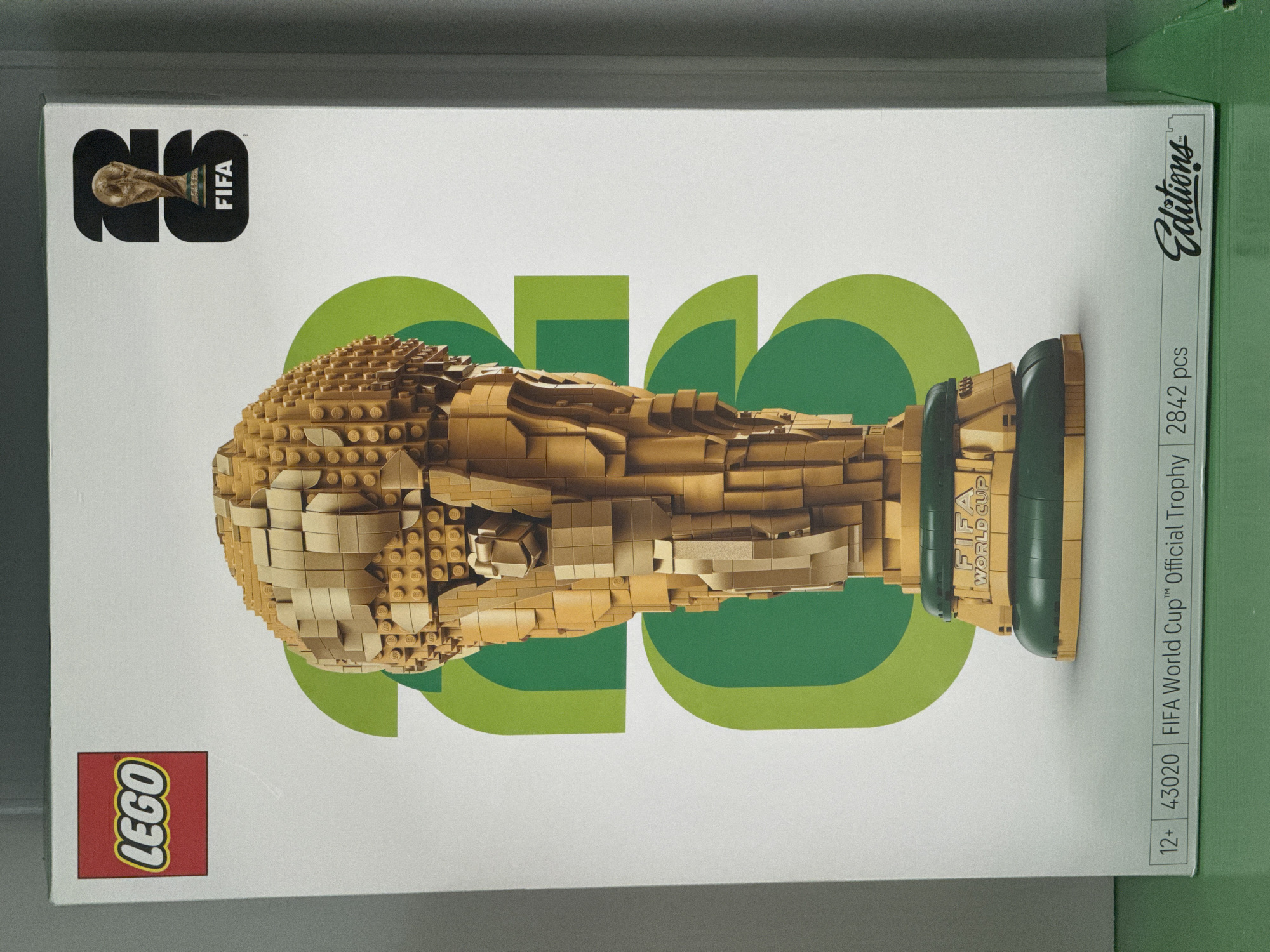 The image shows the front of a LEGO box for the FIFA World Cup Official Trophy set. The box is predominantly white with large green graphic elements in the background. A detailed, golden LEGO replica of the FIFA World Cup trophy is centered on the box. The LEGO logo is visible in the top left corner, and the FIFA logo is in the top right corner, superimposed on a stylized "23". Text at the bottom of the box indicates the set number "43020", the product name "FIFA World Cup™ Official Trophy", and the piece count "2842 pcs". The word "Editions" is also visible in a stylized font in the bottom right corner. The box is placed on a green surface. There is no indication of the city of Castelo Branco, Portugal, in the image.