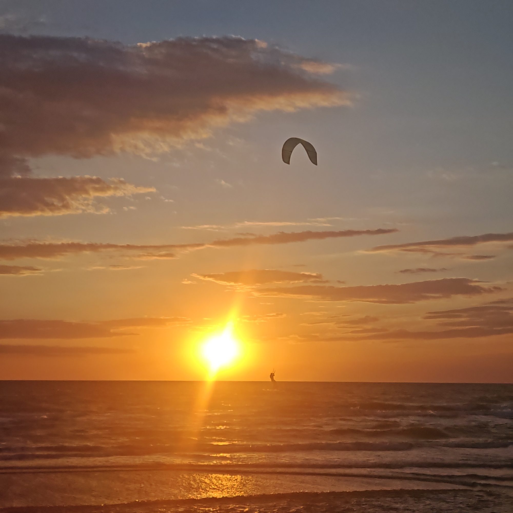 The image depicts a sunset scene at sea, likely in городское поселение Щелкино, Ukraine, based on its coastal location. The sun is setting on the horizon, casting a bright golden glow over the water and sky. A kiteboarder is silhouetted against the sun on the sea, and a kite is visible in the sky. The sky is colored with a mix of orange, yellow, blue, and pink hues, with some clouds scattered around. The sea has gentle waves, and the beach is visible in the foreground.