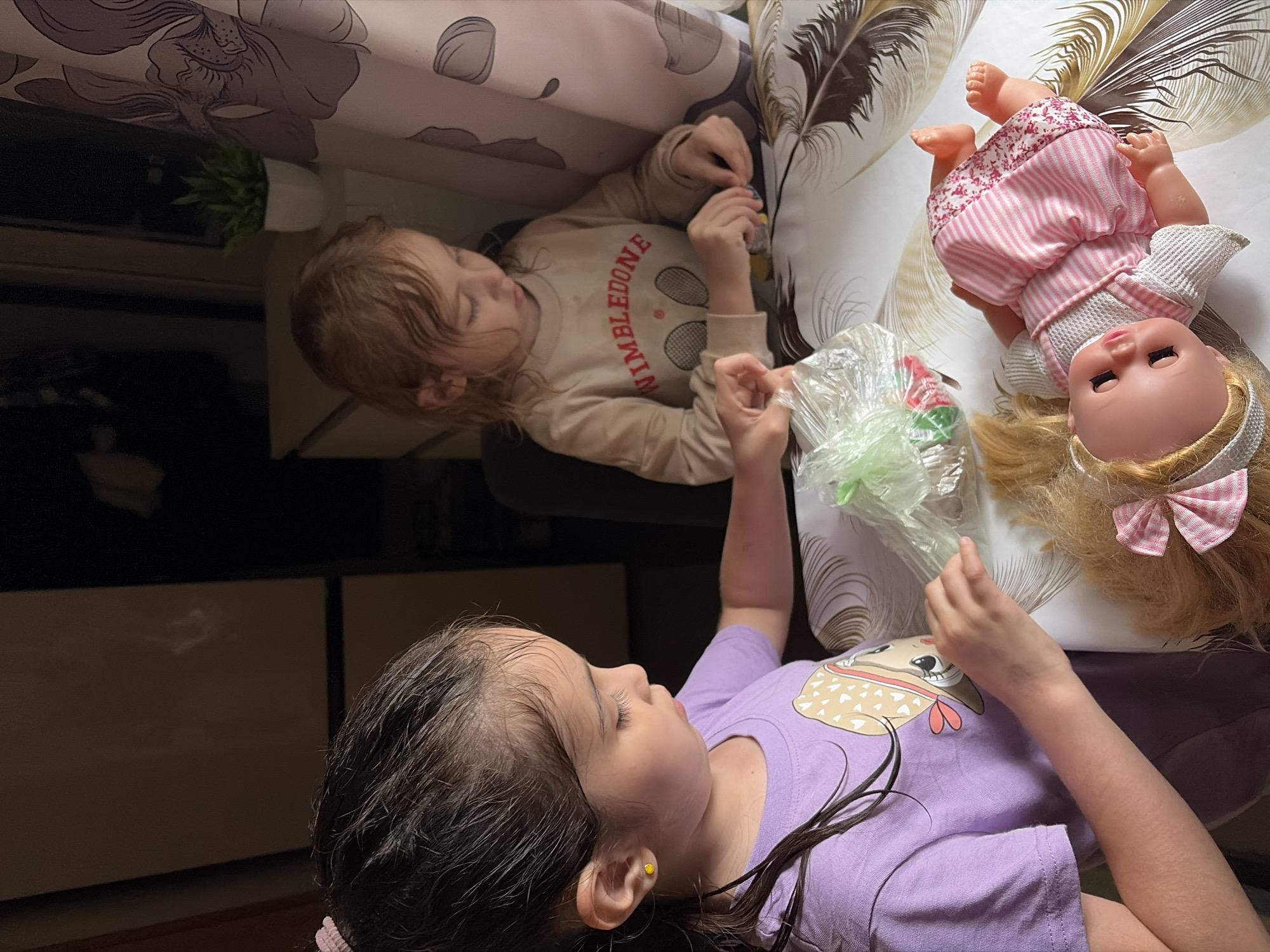 Two young girls are seated at a table. The girl on the left, wearing a purple t-shirt with a cartoon character graphic, is holding a clear plastic bag with green and pink items inside, seemingly engaged in unpacking or playing with the contents. She has dark hair, pulled back, and a yellow stud earring in her left ear. The girl on the right, wearing a beige sweatshirt with "WIMBLEDONE" printed on it and a tennis graphic, is also focused on something on the table, her hands occupied with small objects. Her hair is blonde and she is looking down. In front of the girls lies a blonde doll dressed in a pink and white striped dress with a floral pattern. The table is covered with a tablecloth featuring a feather-like design. The background is dimly lit, suggesting an indoor setting. The overall scene appears to be a domestic environment within the city of Tambov, Russia, likely a home or apartment, given the domestic objects visible such as a plant and curtains in the background, typical of Russian living spaces.