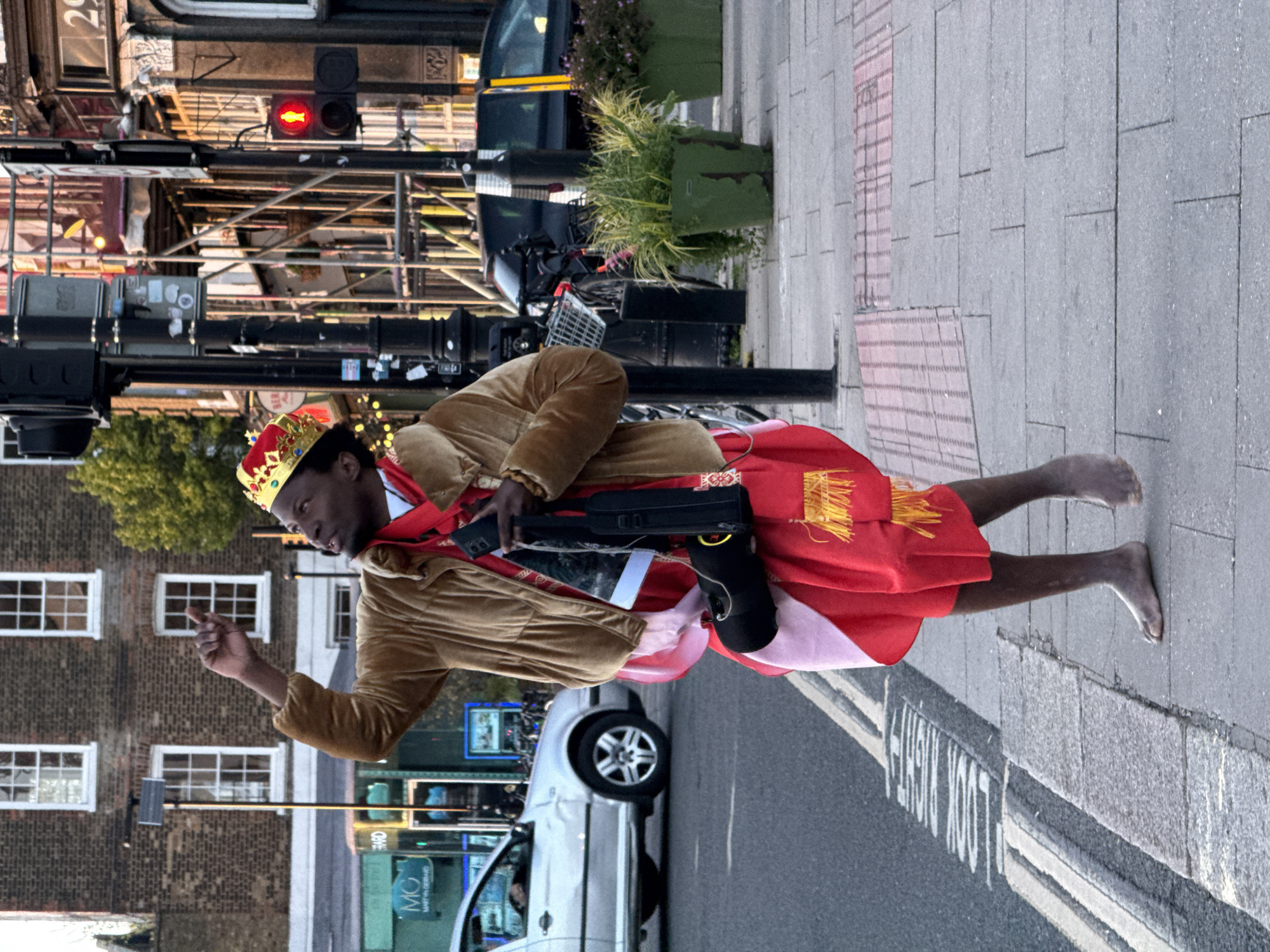 A man wearing a gold crown, a tan jacket, and a red and pink skirt is walking barefoot on a London sidewalk. He is carrying a black case and a smartphone. He is gesturing with his right hand. A white car is passing him on the street. There is a red traffic light in the background, as well as a bike rack and a building under construction. A street sign says "LOOK RIGHT".