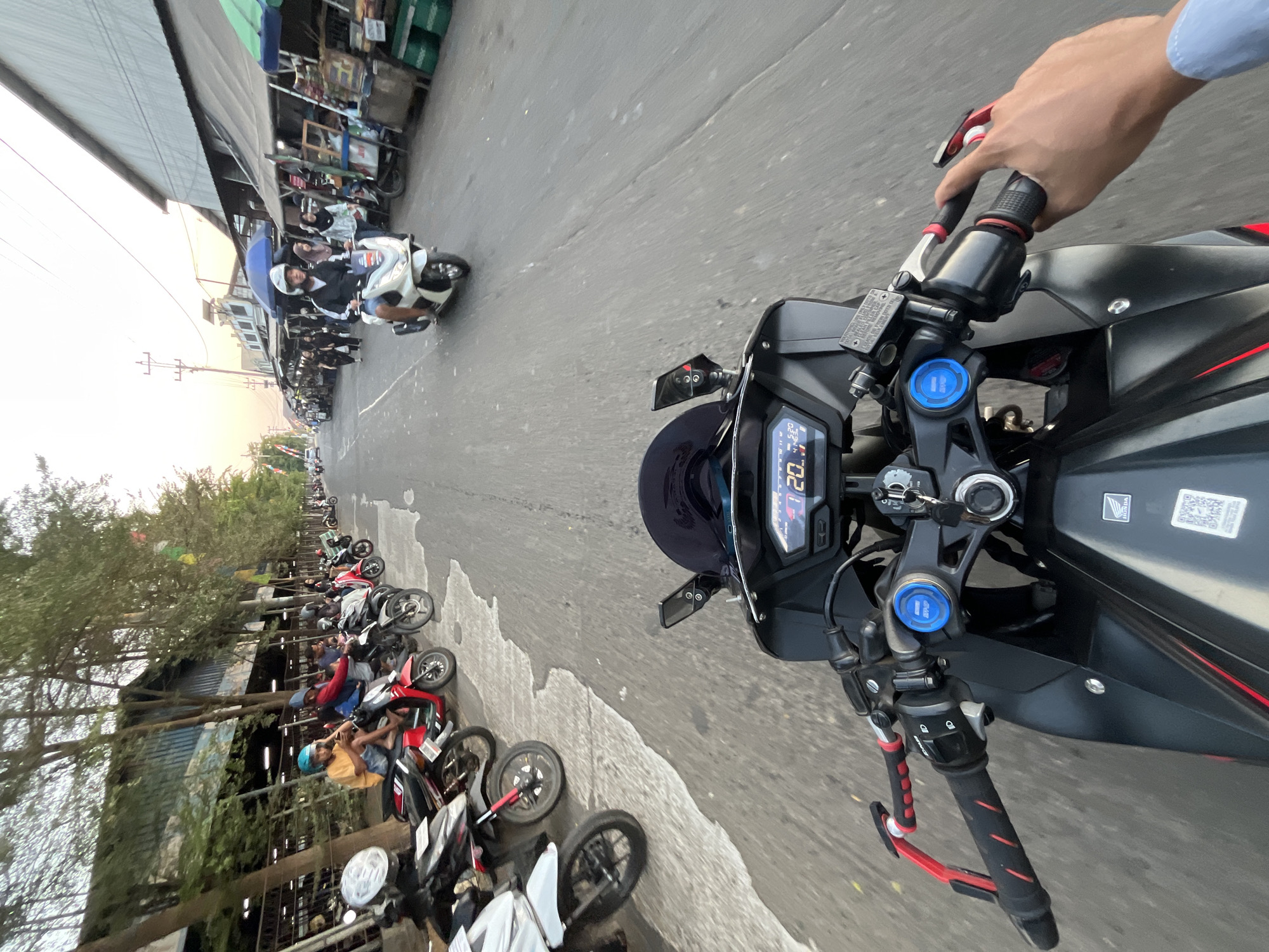 A first-person perspective shot from a motorcycle shows a street scene in Pelang, Indonesia. The road is paved, with parked motorcycles lining the left side. The motorcycle's digital dashboard displays speed as 20.1 km/h and the time as 5:20. On the right side of the road, a row of market stalls with awnings creates a shaded area. Several people are visible, some riding motorcycles and others walking among the stalls. The sky is hazy with the sun low on the horizon, indicating either early morning or late afternoon. The overall atmosphere appears to be a bustling market area during a quiet period of the day.