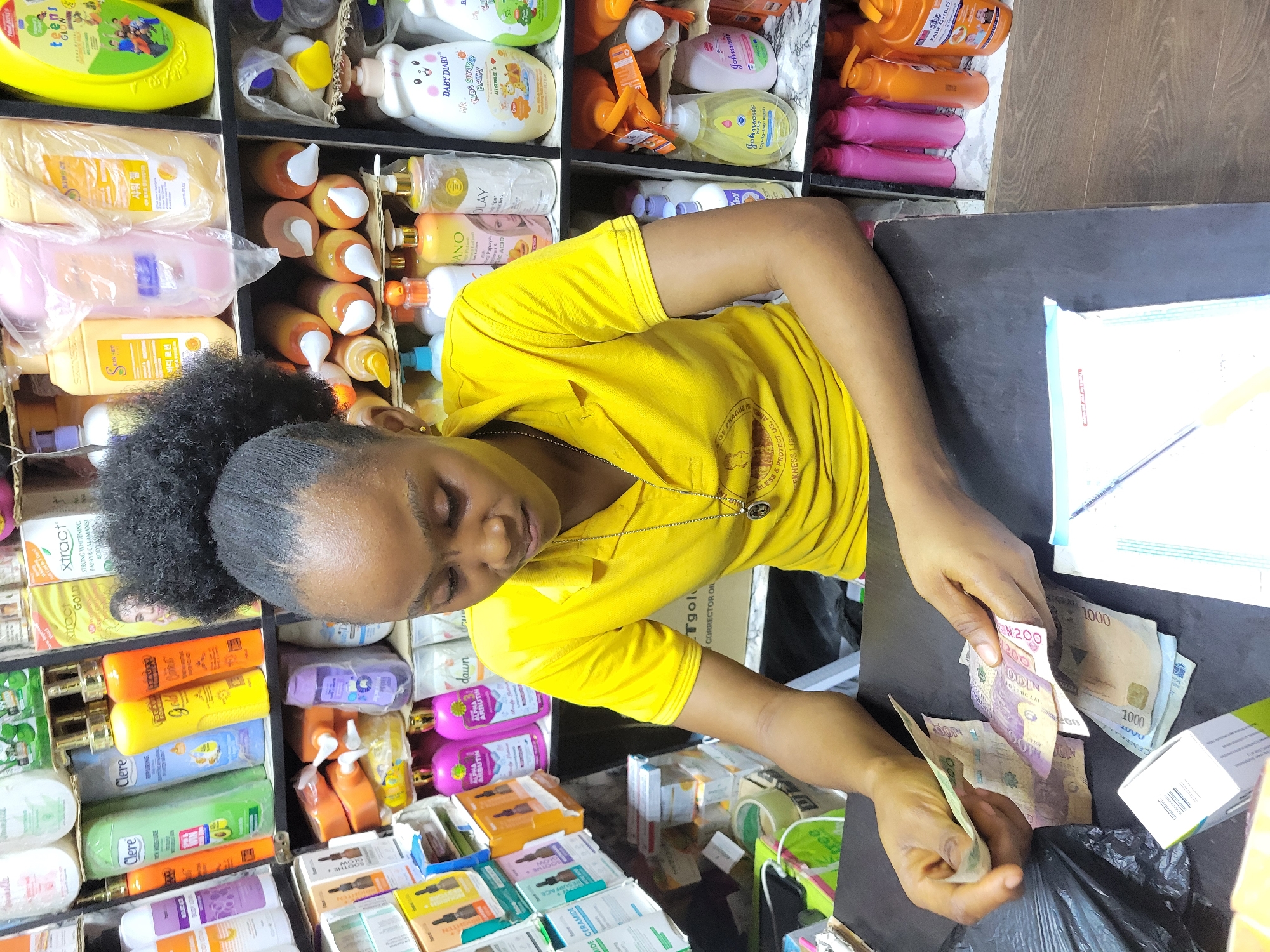 A young woman in a yellow shirt stands behind a counter in what appears to be a small retail shop. Shelves behind her are stocked with various personal care products, including lotions, shampoos, and skincare items. She is counting Nigerian Naira banknotes, specifically ₦200 and ₦1000 denominations. The shop is in Onitsha, Nigeria.