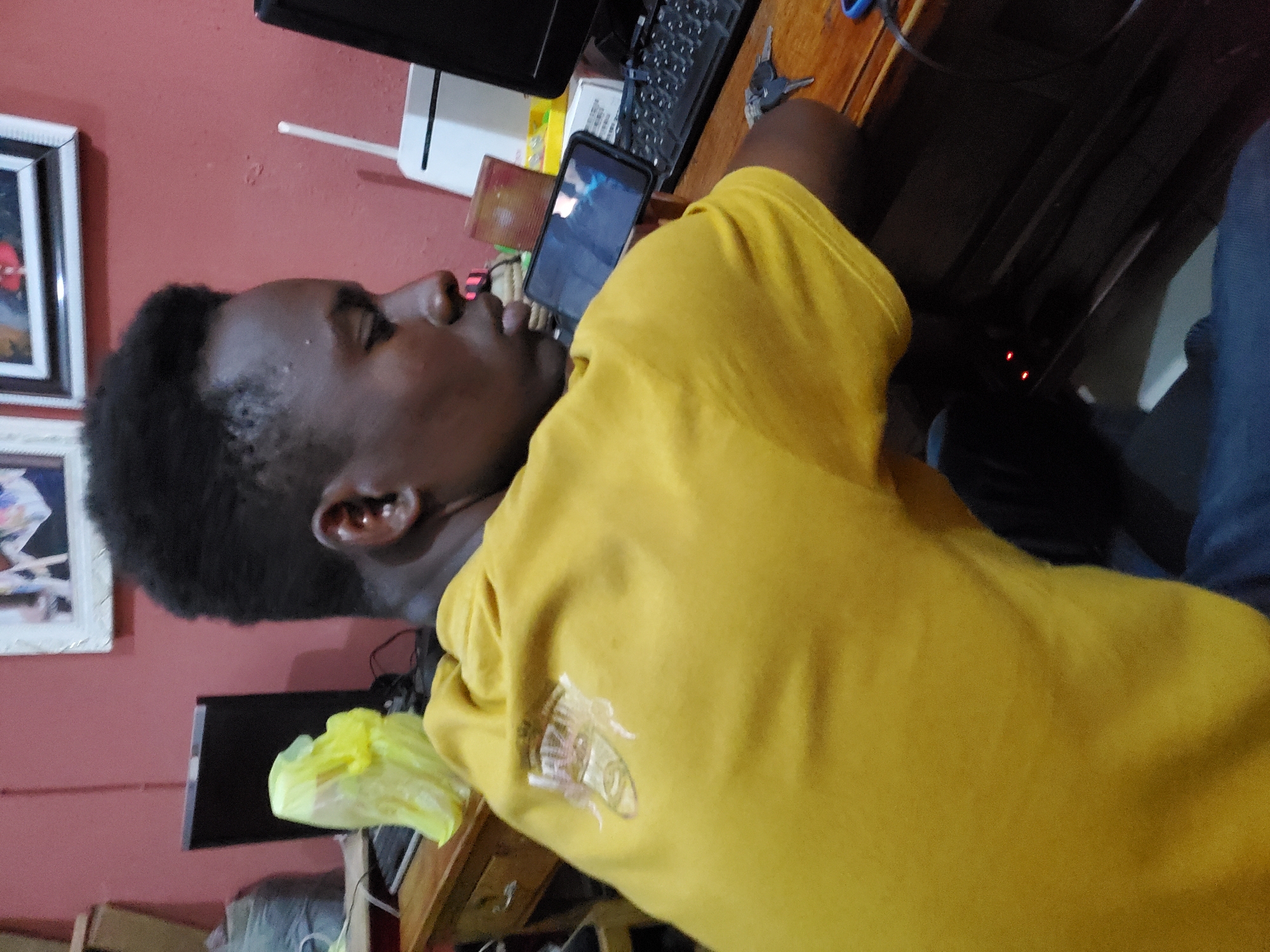 A Black male is reclining on a wooden desk with a mobile phone in hand, possibly located in Mararaba, Nigeria. The phone is displaying a scene on its screen. The desk also contains a black keyboard, computer monitor, a white router, and a yellow plastic bag. The male is wearing a mustard-yellow sweatshirt with a design on the sleeve. The surrounding environment includes red walls and framed pictures.