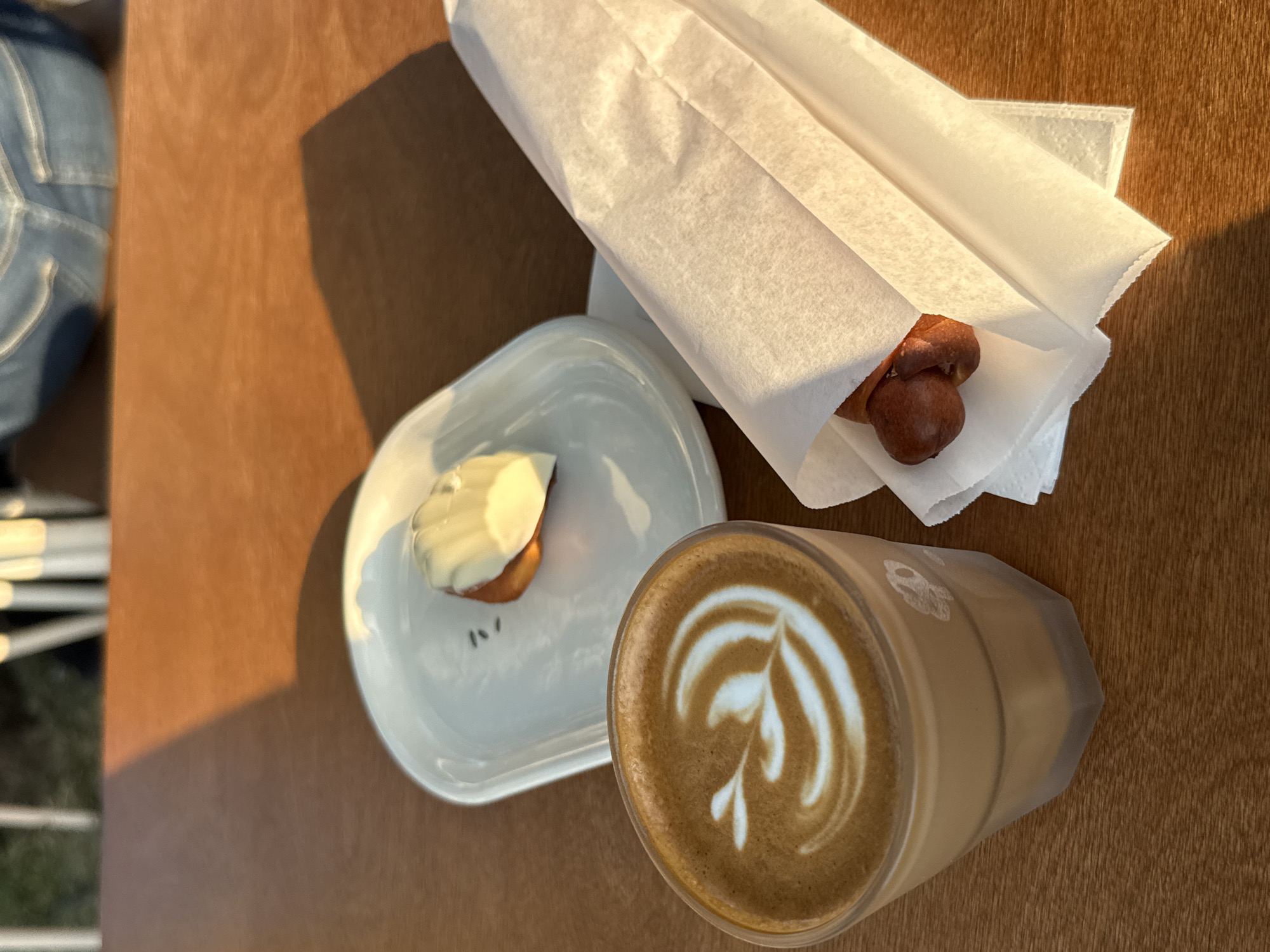 A close-up, eye-level shot shows a coffee drink and pastries on a wooden table in an outdoor setting. The coffee, a latte with latte art, is in a clear glass cup. To the right of the coffee, a pastry is wrapped in white paper. Behind the coffee and pastry, a white plate holds another pastry, possibly a madeleine, topped with white icing. The background is blurred, showing out-of-focus vertical lines and the suggestion of people wearing denim jeans. Sunlight casts shadows across the table. The scene appears to be a casual outdoor cafe or dining area.