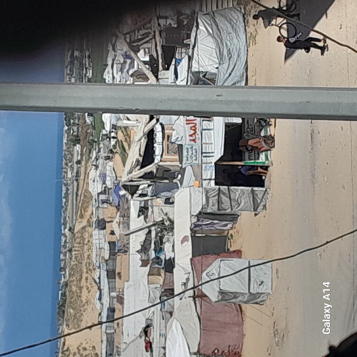This image presents an elevated, wide view of a densely populated, makeshift settlement situated in Khan Yunis, Palestinian Territory, under bright daylight. A light-colored dirt road or path traverses the mid-ground. On the right side of this road, two individuals are visible: one person in dark clothing walks rightward, holding a dark object, adjacent to a black bicycle; another individual is partially obscured while seated on a bicycle further to the right. To the left of a prominent vertical gray pole that partially obscures the right foreground, a small structure or shop with a white canopy displays Arabic signage, reading "المجد" (Al-Majd) and additional text. Two people are near this structure's entrance; one in a blue shirt is standing inside, while another appears to be working near a cart outside. The mid-ground is filled with numerous improvised shelters and tents of varying sizes, predominantly white, brown, and red-brown, constructed from tarpaulins, canvas, and salvaged materials. One reddish-brown tent has a red circular emblem on its side. Beyond the settlement, barren sandy hills extend towards the horizon, where a distinct strip of the Mediterranean Sea is visible under a clear blue sky with light clouds. The visible terrain is predominantly sandy and dusty.