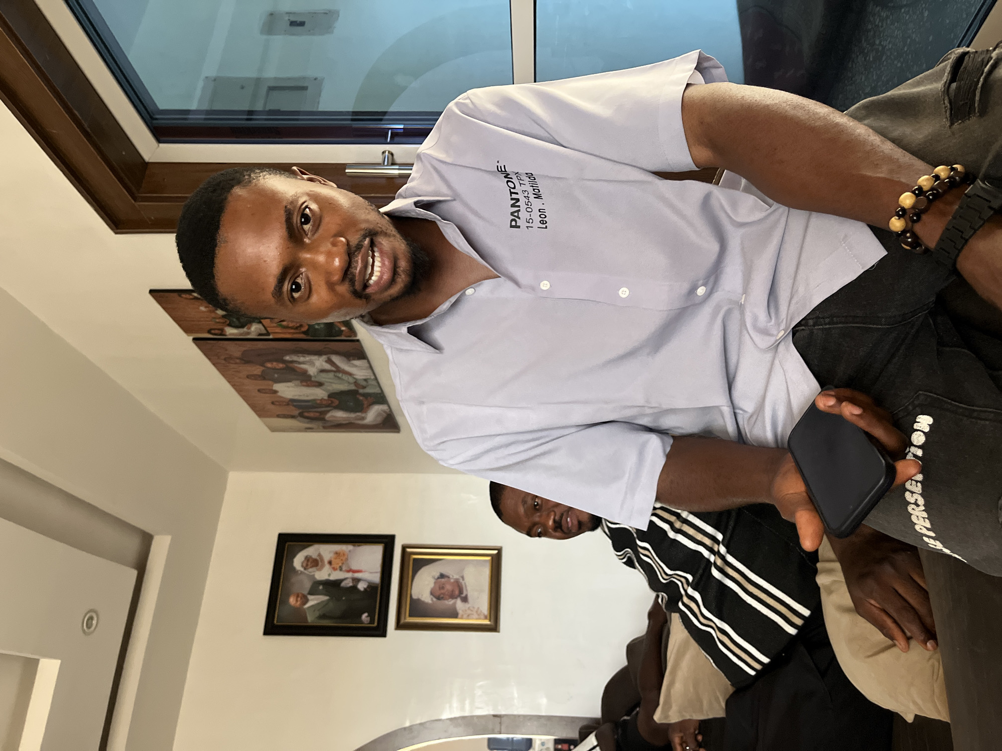 The image, rotated 90 degrees clockwise, captures two men indoors in what appears to be a living room or common area in Jos, Nigeria.
The primary subject, a man with dark skin, short dark hair, and a beard, is positioned in the foreground. He is wearing a light grey short-sleeved button-down shirt and dark jeans. His left wrist is adorned with a beaded bracelet and a dark-colored watch. He holds a black smartphone in his right hand. He is looking directly at the camera with a broad, friendly smile, indicating awareness of being photographed.
On his shirt, visible text reads: "PANTONE", "15-0543 TPX", and "Leon . Matilda". His dark jeans feature partially visible white text along the outer leg, which appears to spell out "PERSEVERANCE" or a similar word.
To the right and slightly behind the main subject, a second man with dark skin is partially visible. He is wearing a black and white striped shirt.
The background shows a brightly lit room. A large window with a dark frame is prominent in the upper left, letting in natural light. On the wall, several framed pictures are displayed. Two are clearly discernible: one depicting a wedding couple (man in a dark suit, woman in a white dress), and another, smaller picture in a gold frame, showing a woman in a white head covering. A larger, more ornate picture with multiple figures is also partially visible above these. The overall impression is one of casual comfort and a friendly atmosphere during the daytime.