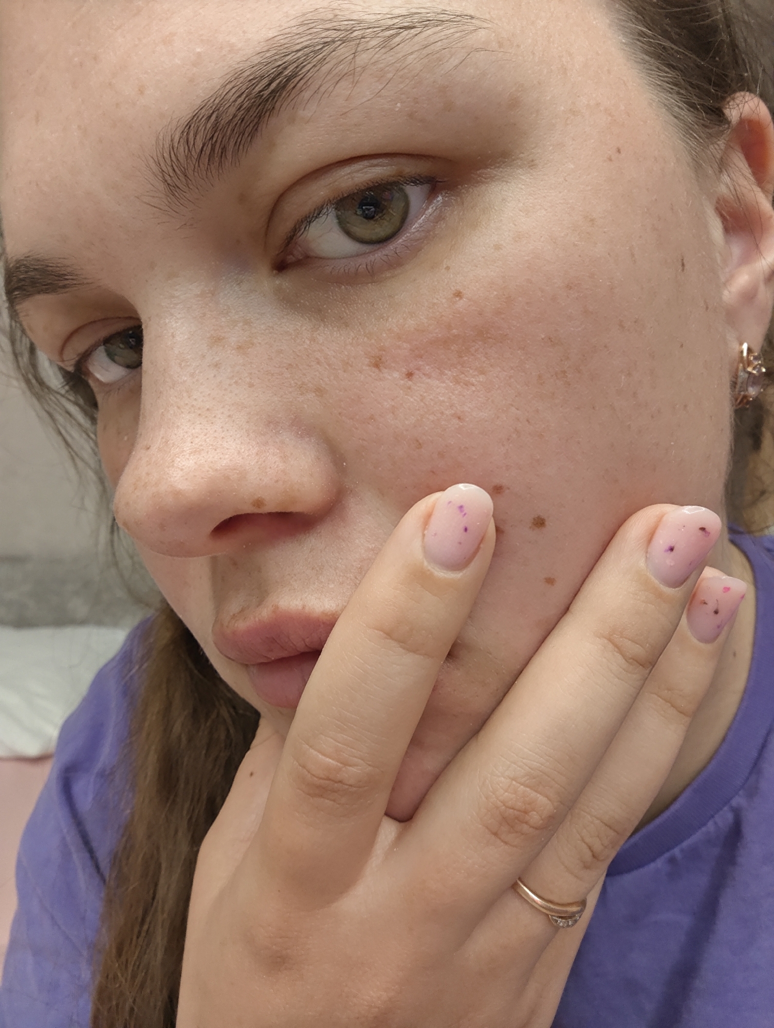 A close-up, indoor shot depicts a Caucasian woman with fair skin, freckles, and hazel eyes. She is wearing a light purple t-shirt. Her brown hair is pulled back. She has a gold ring on her ring finger and gold earrings. Her nails are manicured with a light pink polish with small darker flecks. Her hand is touching her face. The background appears to be a blurred room with a bed. There is no contextual information to identify the location as the city of Chaikovsky, Russia.