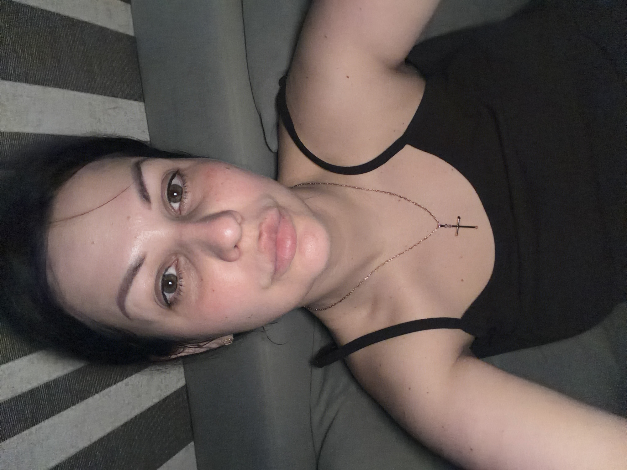 A young adult woman is depicted in a close-up, casual shot, lying on her back on a grey upholstered surface, likely a sofa or bed. Her head is positioned towards the left of the frame, and her body extends towards the right. She has dark hair, dark eyebrows, and brown eyes that are looking directly at the camera. Her lips are slightly puckered in a playful or "kissy face" expression. Her fair skin shows a natural flush or redness on her cheeks. She is wearing a dark (likely black) tank top or camisole with thin straps. A delicate gold-toned chain necklace with a small cross pendant rests on her chest. The background consists of the solid grey upholstery and a striped pattern of varying shades of grey, possibly part of a cushion or blanket. The lighting is dim, indicating an indoor setting, possibly in the evening or at night. The overall impression is one of relaxed informality. There is no visible text in the image. The location context for this image is Sochi, Russia.