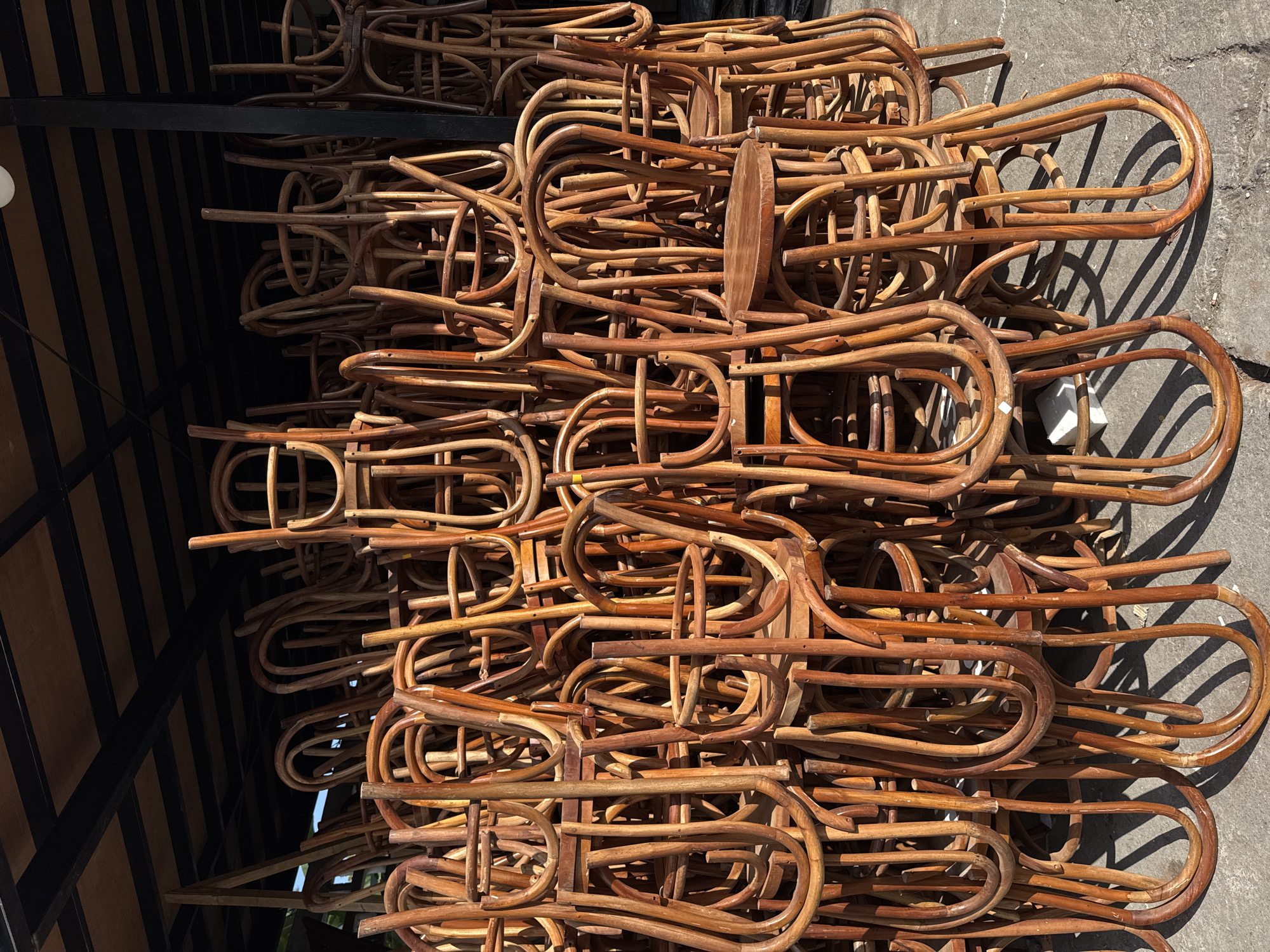 A large, outdoor stack of bentwood chairs sits beneath a dark, slatted canopy structure in North Kuta, Indonesia. The chairs are made of a light brown wood with a visible grain. Their curved backs and legs form a dense, repeating pattern, with many chairs nested inside one another. The ground beneath the chairs is a rough, grey concrete surface, with shadows cast by the chairs and the overhead structure. The scene appears to be a storage area for a restaurant or cafe, with the chairs piled high and waiting to be used. The lighting suggests it is daytime, with sunlight filtering through the gaps in the canopy, creating areas of light and shadow. There are no people visible in the image.