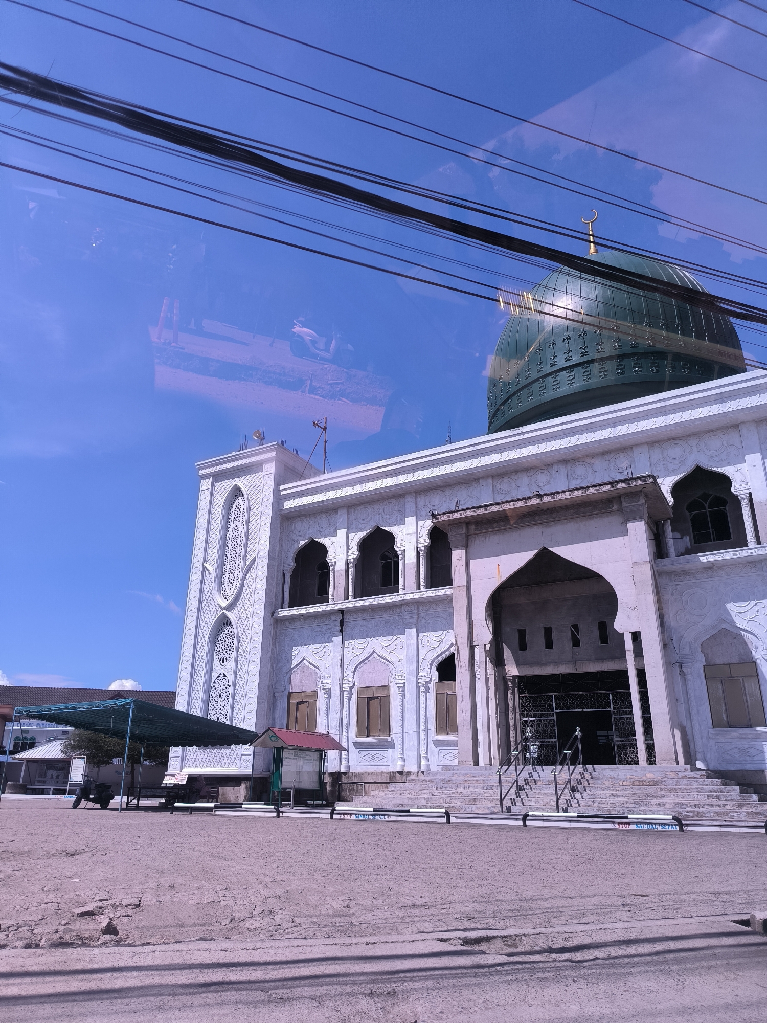 The image shows a large, white mosque with a prominent green dome, captured on a clear, sunny day. The photograph appears to be taken through a glass surface, as evidenced by reflections of overhead power lines, clouds, and faint shapes that might include a motorcycle and other elements of an outdoor scene, particularly visible in the upper and lower left portions of the image.
The mosque itself is a striking white building adorned with intricate geometric carvings and arched windows. A tall section on the left side of the mosque, possibly a minaret or a decorative tower, also features these detailed patterns. The main entrance is grand, with a large arched doorway leading to steps with metal railings. The green dome is topped with a golden crescent moon finial and features decorative bands.
In the foreground, there is an unpaved, dusty or gravelly area leading up to the mosque's steps. To the left of the mosque, a green canopy provides shade, and a small, red-roofed kiosk or shelter is also visible. Near the base of the mosque steps, low barriers are in place, one of which clearly displays the text "STOP SANDAL SEPATU" (Stop Sandals Shoes).
The weather is bright and sunny, indicated by the clear blue sky and strong shadows. No people are explicitly visible in the main scene. The image captures the mosque's architectural beauty within its immediate surroundings in Keudelapang, Indonesia.