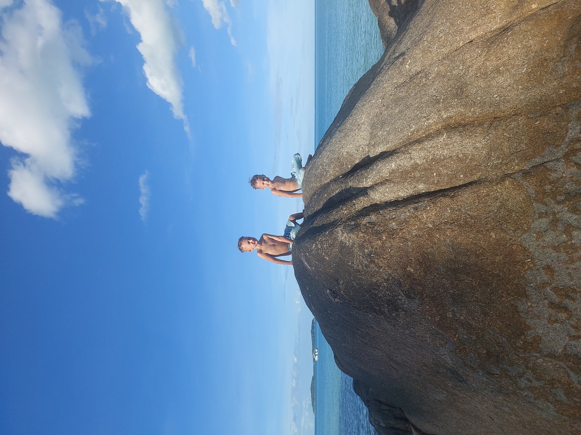 Two young Caucasian boys are sitting on a large, rounded boulder on a beach. The boy on the left is looking to his left with his mouth open. The boy on the right is looking forward. Both boys are shirtless and wearing light-colored swim trunks. The boulder is dark brown and has visible texture. In the background, the ocean is a calm, turquoise color, and the sky is a bright blue with scattered white clouds. In the distance, there is a small island or landmass and a boat. The scene appears to be set in Baan Chaweng Noi, Thailand.
