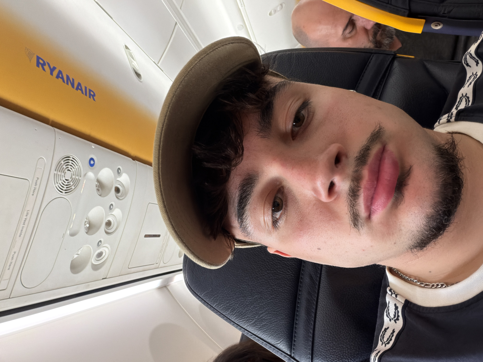 A close-up photograph taken inside a Ryanair airplane. The primary subject is a young man wearing a tan hat, black t-shirt and a silver chain, seated in a black leather airplane seat, angled slightly to the right. Behind him and above his head is another passenger. Above and to the left of the young man are the overhead air vents and reading lights. A panel with the Ryanair logo is visible above the fixtures. The photograph appears to have been taken by the young man himself. There is no indication of a location in the city of Matosinhos, Portugal, or any other specific location.