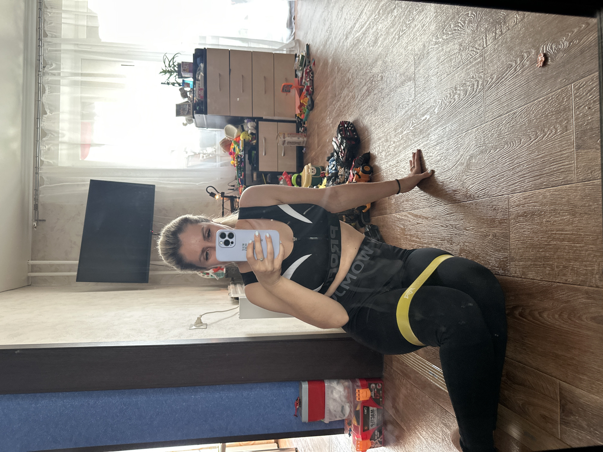 A woman is sitting on a wooden floor in what appears to be a living room. She is wearing a black sports bra and black leggings with a yellow resistance band around her thighs. She is holding a phone in her right hand, taking a selfie. The room is cluttered with toys scattered on the floor and a dresser with several drawers. There is a window with white curtains in the background. The woman's hair is blonde and pulled back. The image is taken in Novoaltaysk, Russia.