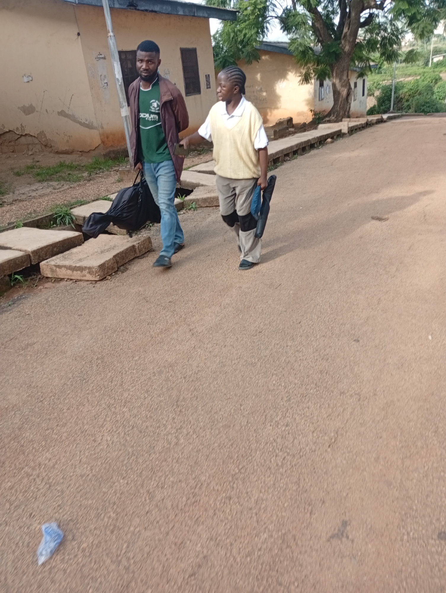 Two individuals, a man and a youth, walk on a paved road in what appears to be Rafin Par, Nigeria. The man, on the left, wears a green t-shirt under a brown jacket and blue jeans, carrying a large black bag. The youth, on the right, is dressed in a yellow vest over a white shirt and patterned shorts, holding a dark object. They are walking away from the camera, towards a tree-lined area with buildings in the background. The road surface is sandy and textured, with concrete slabs lining the left side. The environment suggests a rural or semi-urban setting, with a building exhibiting signs of wear and tear. The lighting indicates it is daytime, with shadows cast on the road.