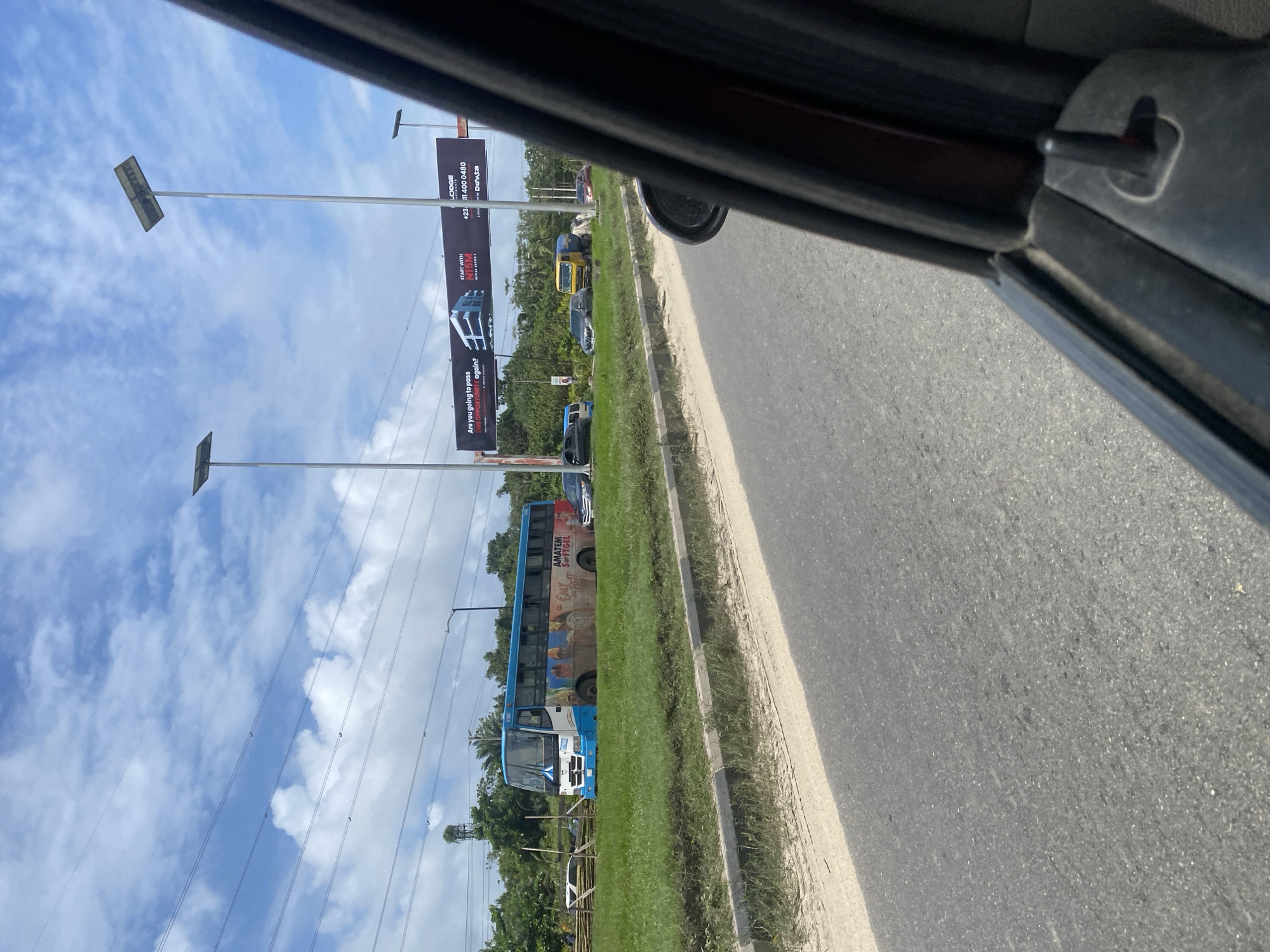 The image captures a street view from inside a vehicle on a bright, partly cloudy day, likely in Lekki Peninsula II, Nigeria. The perspective shows the interior of a car, including the side view mirror and a portion of the door frame. Outside, a paved road runs parallel to a grassy median strip. On the median, a blue and white bus with colorful advertisements is parked. Further down the road, other vehicles, including a yellow truck and a dark sedan, are visible. A large billboard advertising apartments is also present. The background features lush green vegetation and trees. Several tall streetlights with solar panels are spaced along the roadside. A few people can be seen in the distance near some wooden structures.